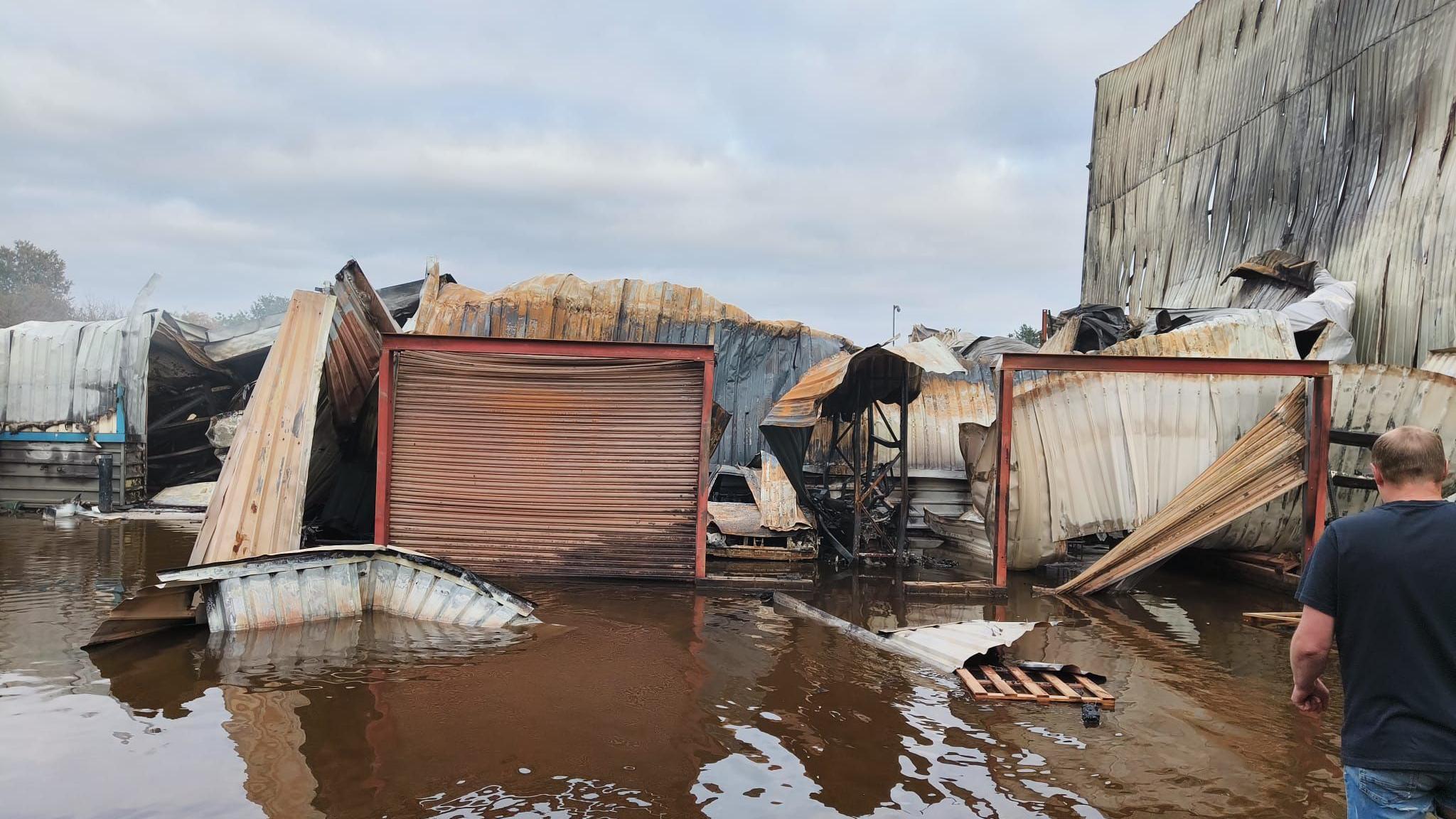 The remains of a building that has been flooded following a fire. Scraps of metal are collapsed around what were a pair of garage doors. Brown water is on the ground. The back of a man wearing a blue T-shirt is at the right of the frame. 