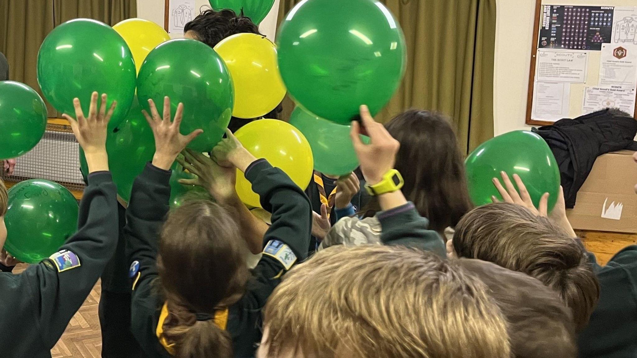 Children in green Scout uniforms have their arms in the air and are holding up green and yellow balloons