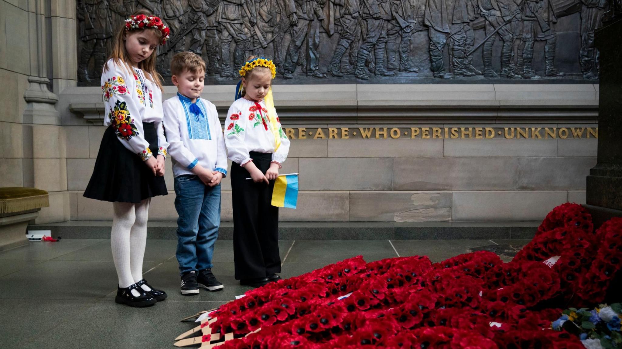 Three young Ukrainian children look down at poppy wreaths at a war memorial. They are wearing Ukrainian national dress. Two girls are wearing flower garlands in their hair and one is holding a small Ukrainian flag  