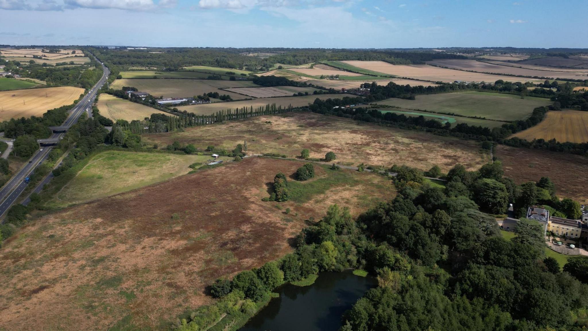 A drone view shows the proposed site of the studio, which is open landscape with a road on the left of the picture.