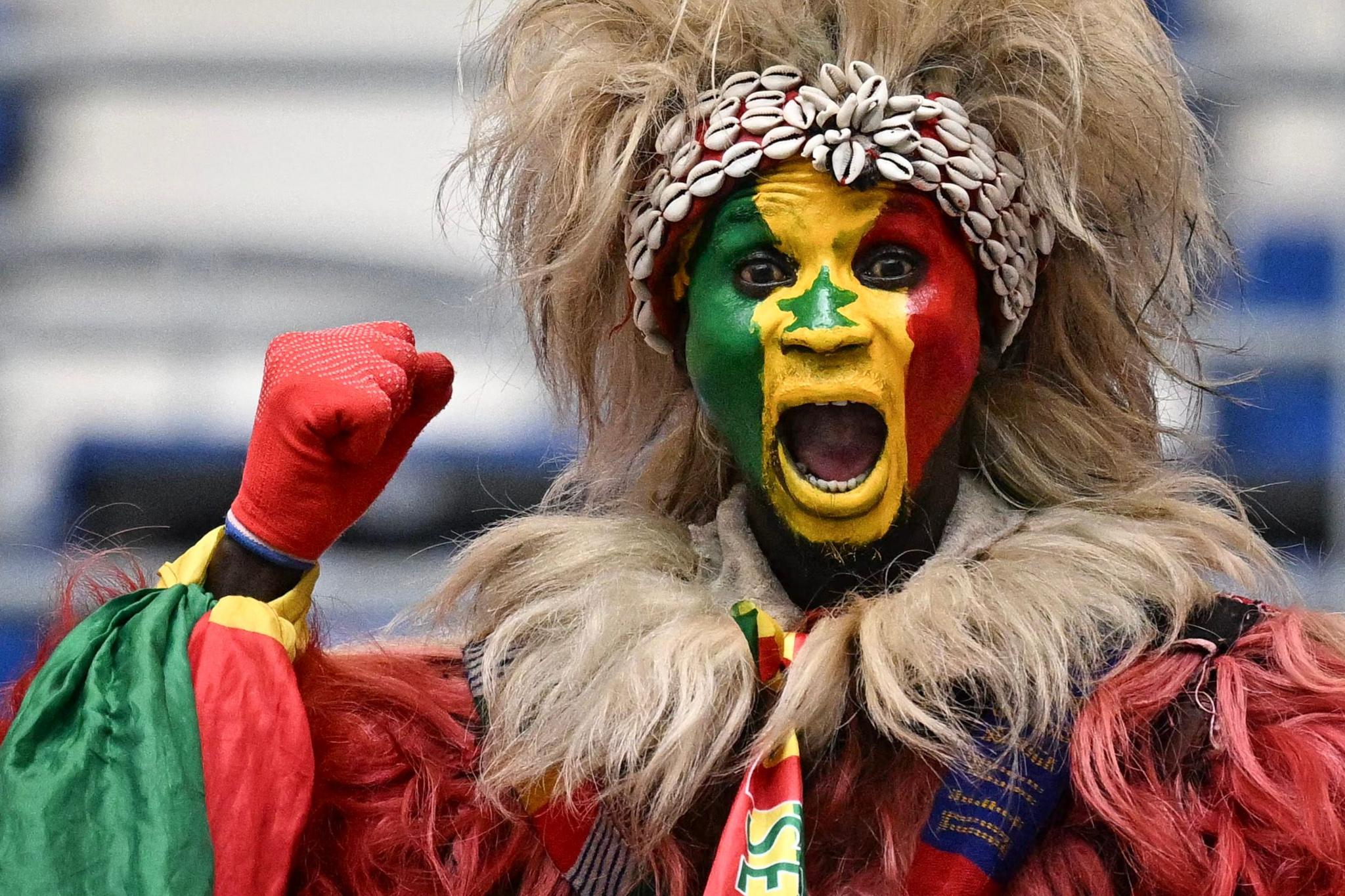 A supporter wearing green, yellow and red face paint raises a clenched fist and shouts.
