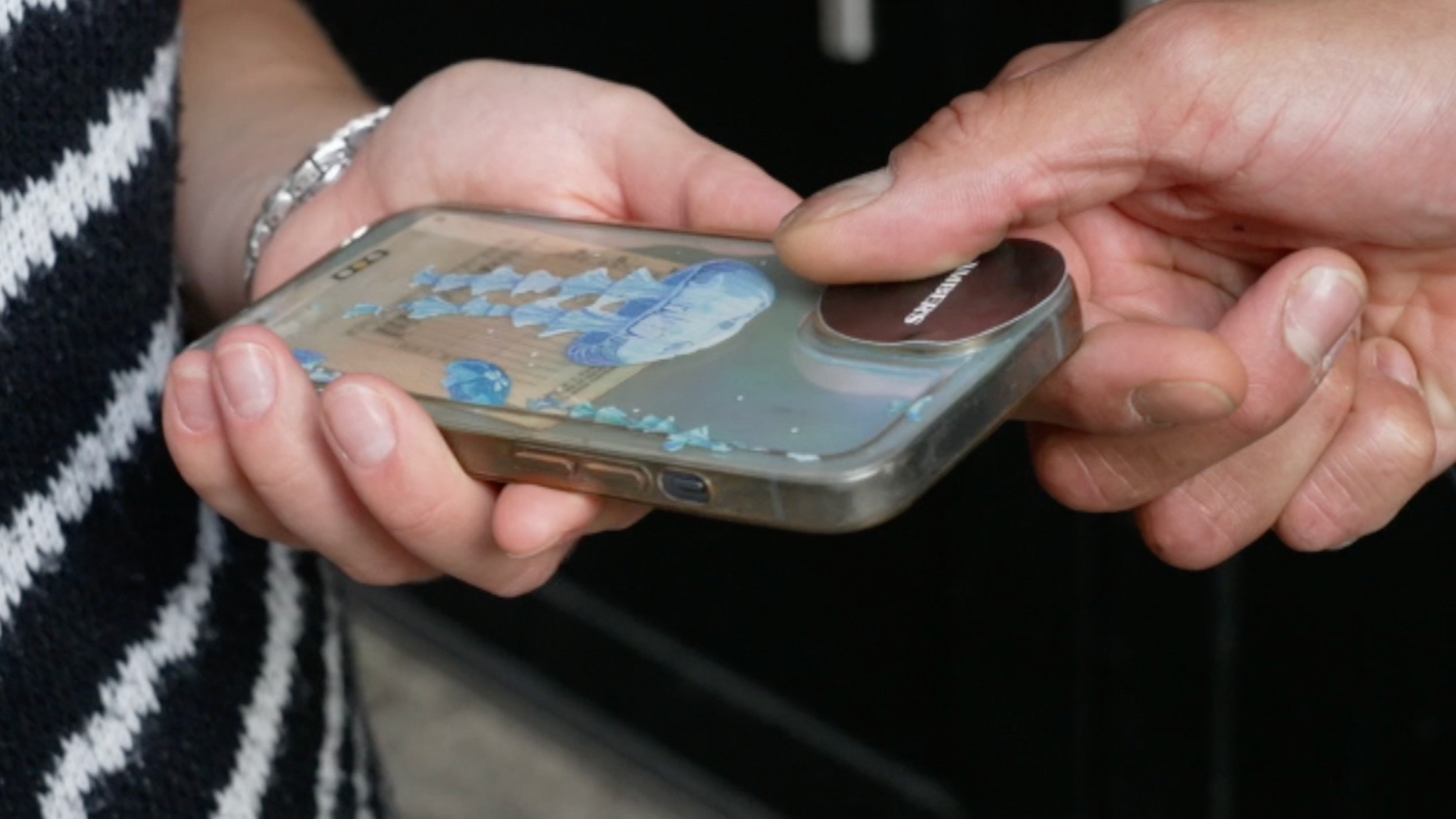 One hand, holding a phone, with another hand reaching out to put a sticker over the phone camera