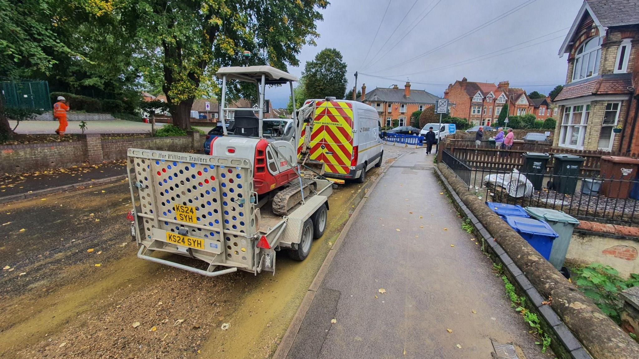 Road with homes along the right-hand side. The road is covered in debris from flooding a Thames Water van and trailer with digger is parked up on the road.