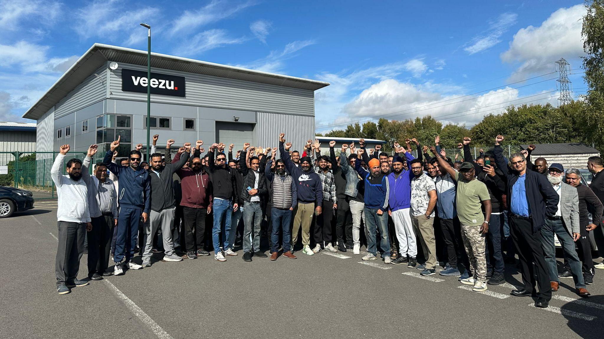 A group of several dozen men standing in a car park in front of a building with a sign reading "Veezu". They are standing in a group and have their hands raised in the air with fists up.