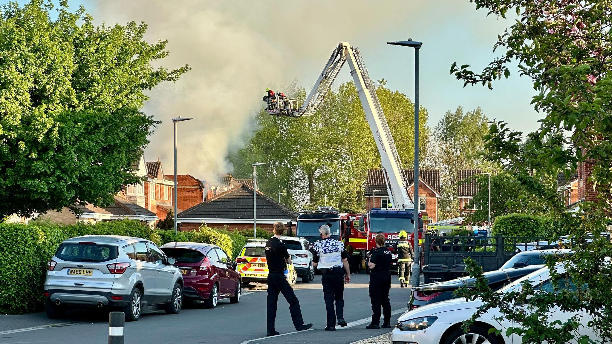 Three people stand in the street as thet watch a fire engine with an aerial platform working to extinguish a fire. A large plume of smoke can be seen above some houses.