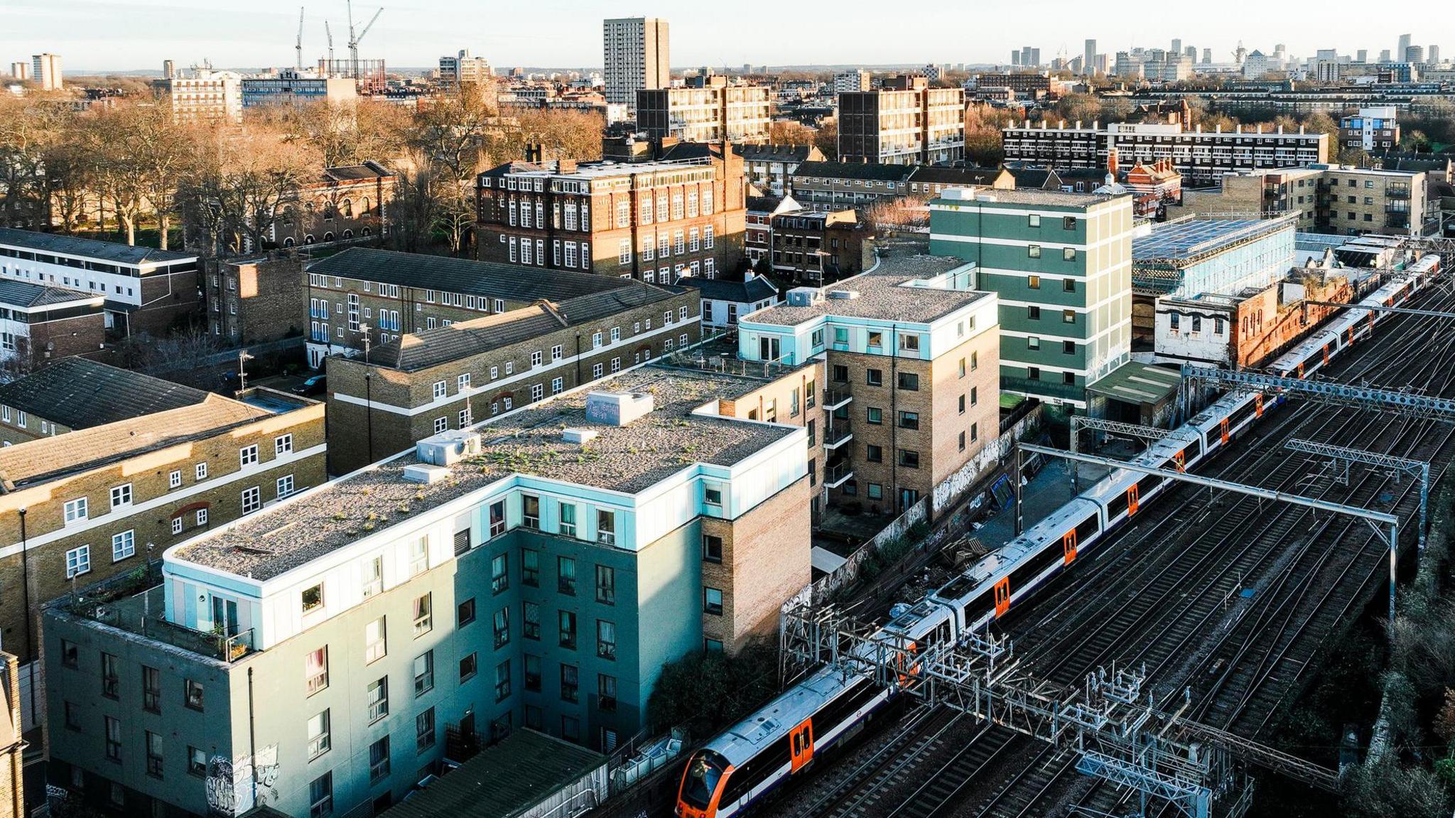 A train line runs past a residential area in London, with the London skyline in the background. The residential area mostly consists of low-rise blocks of flats. A train is running on the train tracks past the flats.