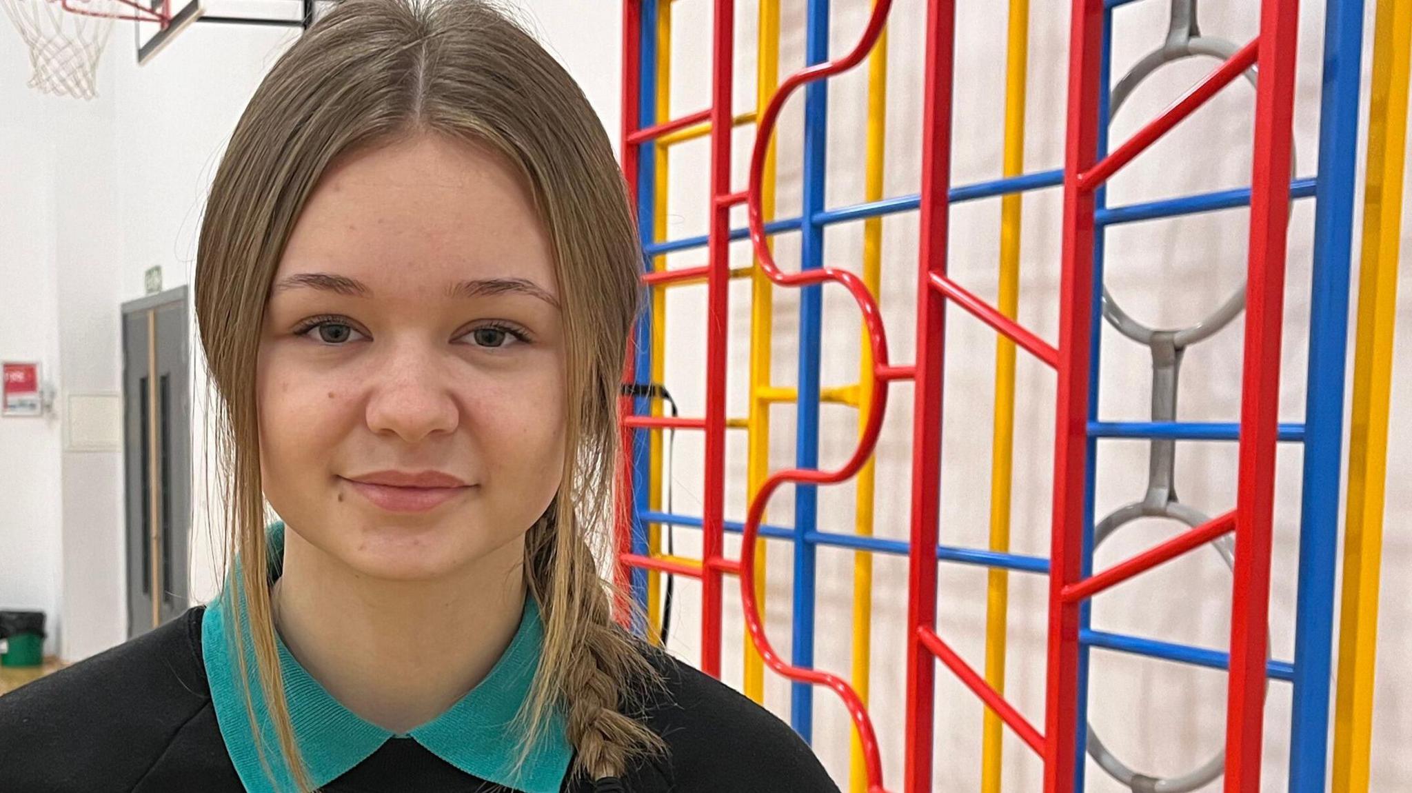 Lily-Kim stands in a school gym beside climbing bars coloured red, blue and yellow. She has long light-brown hair, tied in a plait, and wears an aquamarine polo shirt and a black pullover. 