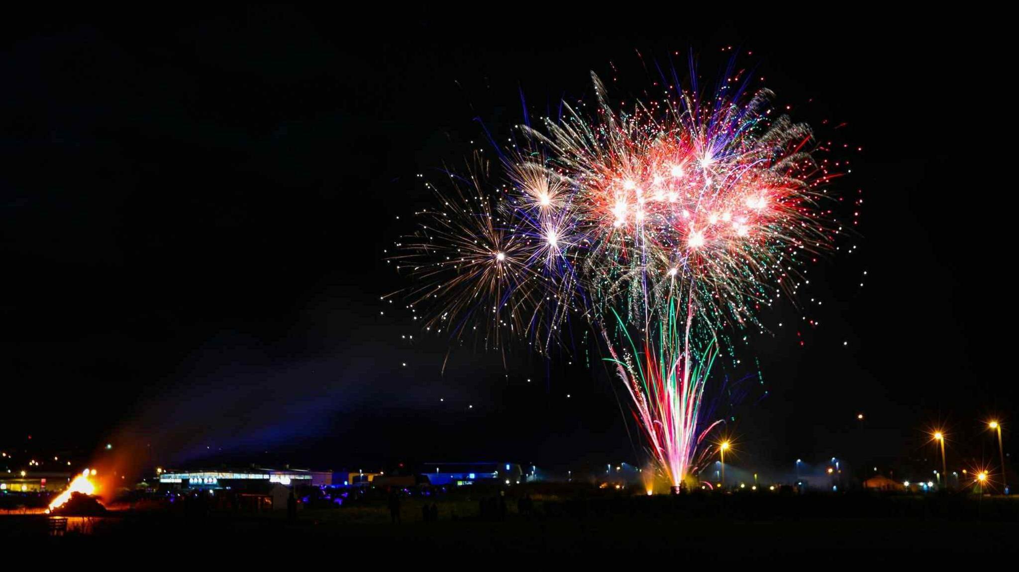 Colourful fireworks against a dark night sky. A bonfire burns in the bottom left corner.
