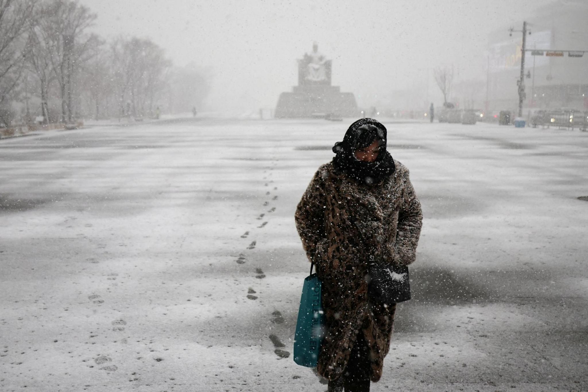 A woman wrapped up for winter weather in a thick coat and scarf around her head walks through a deserted street as snow falls and blankets the ground. There is a monument in the distance behind her.