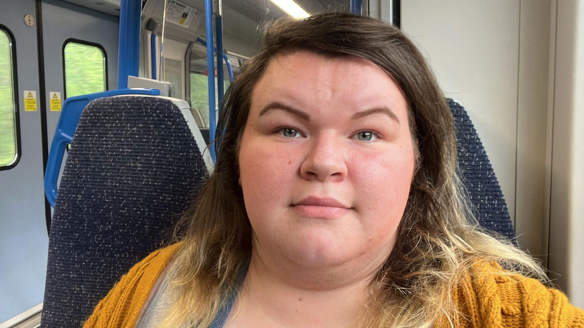 Nikki, who has brown hair and is wearing a knitted yellow cardigan, sits in a train carriage with an empty seat beside her. She looks directly at the camera, face neutral.