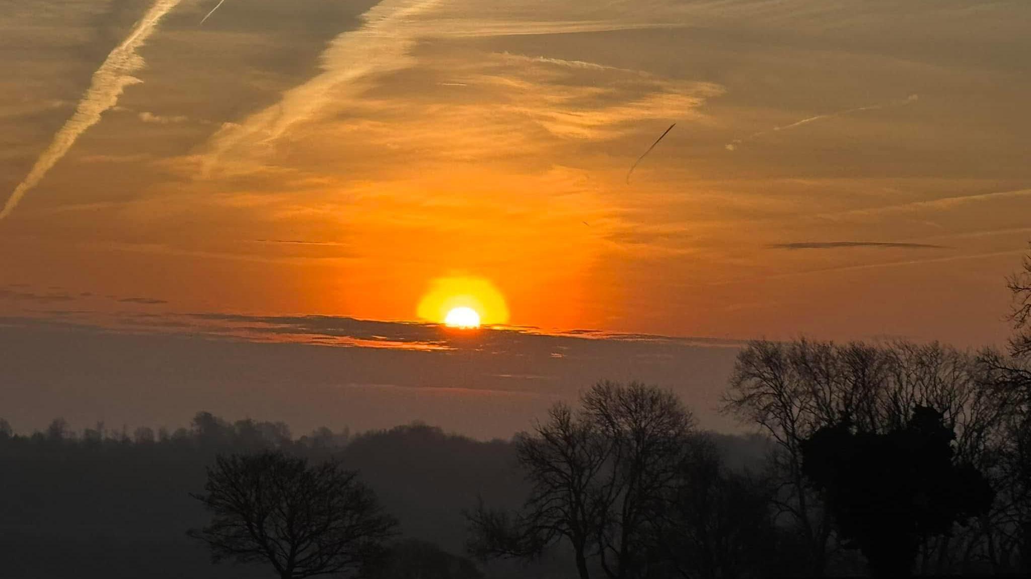 Sunrise with an orange sky and orange halo around the sun. There are trees and a small range of hills silhouetted in the foreground