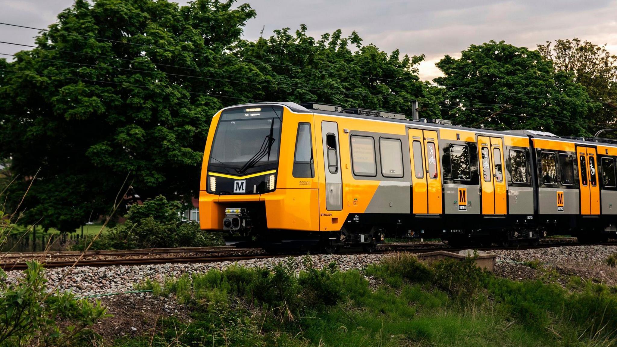 A new yellow, grey and black Metro train that is travelling along a track. The track is above an embankment in the foreground, while behind it are tall trees.