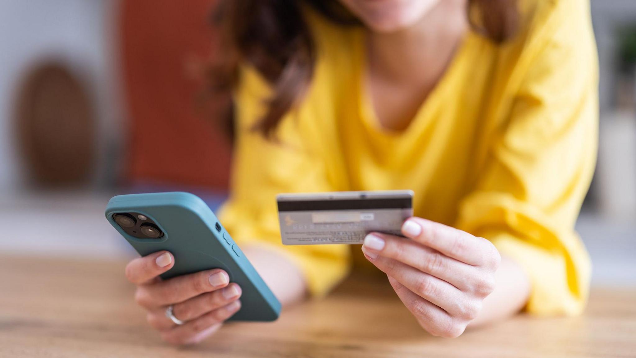 A woman wearing a bright yellow jumper, whose face cannot be seen, handles a smartphone in one hand and a credit card in another while leaning on a kitchen worktop.