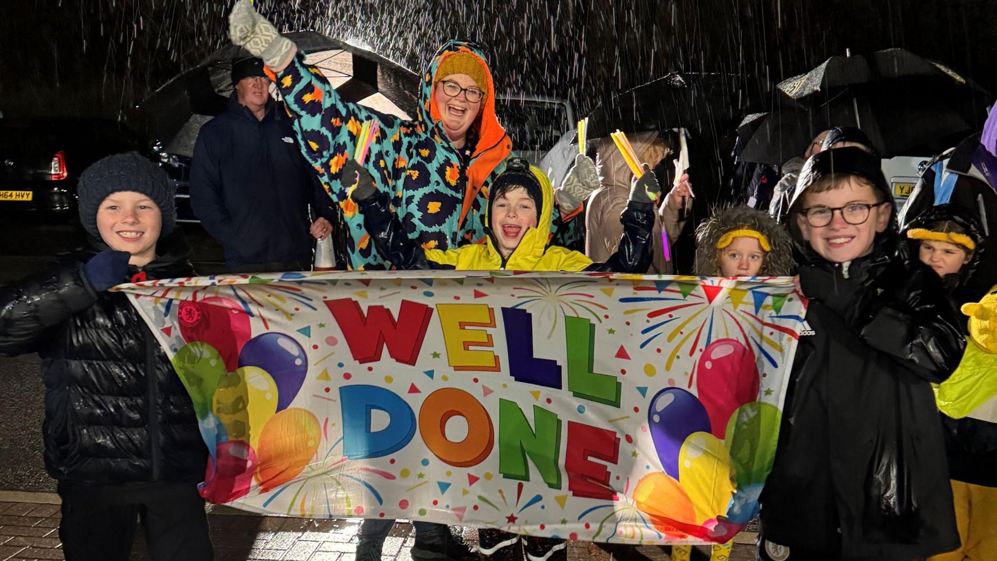 Four children and a woman stood outside with smiles on their faces. The children are holding a colourful banner with balloons on which reads 'Well done'.