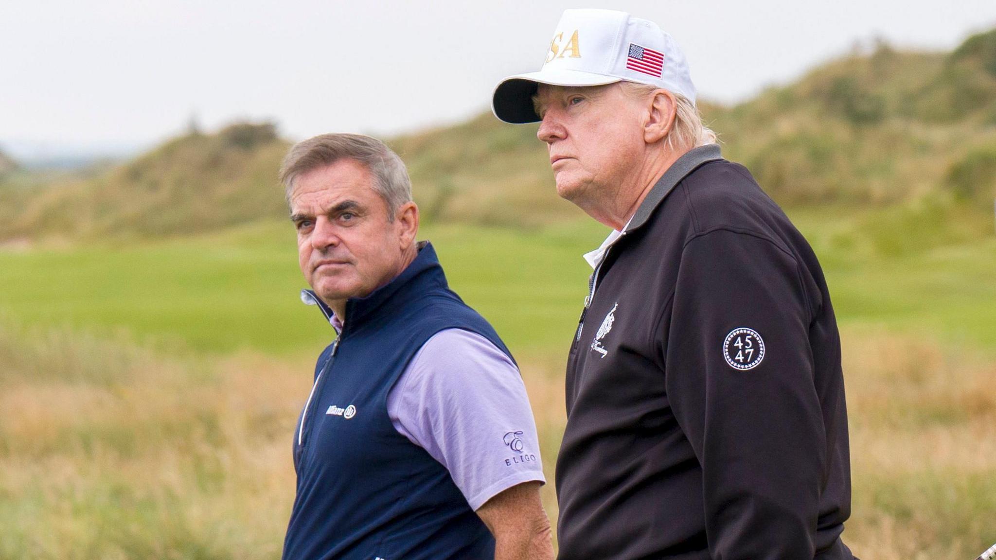 Two men walk along the fairways of a golf course. President Trump is wearing a black half zip windcheater and a white baseball cap, with USA in gold letters. It also has a saltire on the side. Paul McGinley has brown/ grey hair combed in a side shed . He is wearing a purple polo shirt and a sleeveless blue half zip. Both men are looking off to the left.
