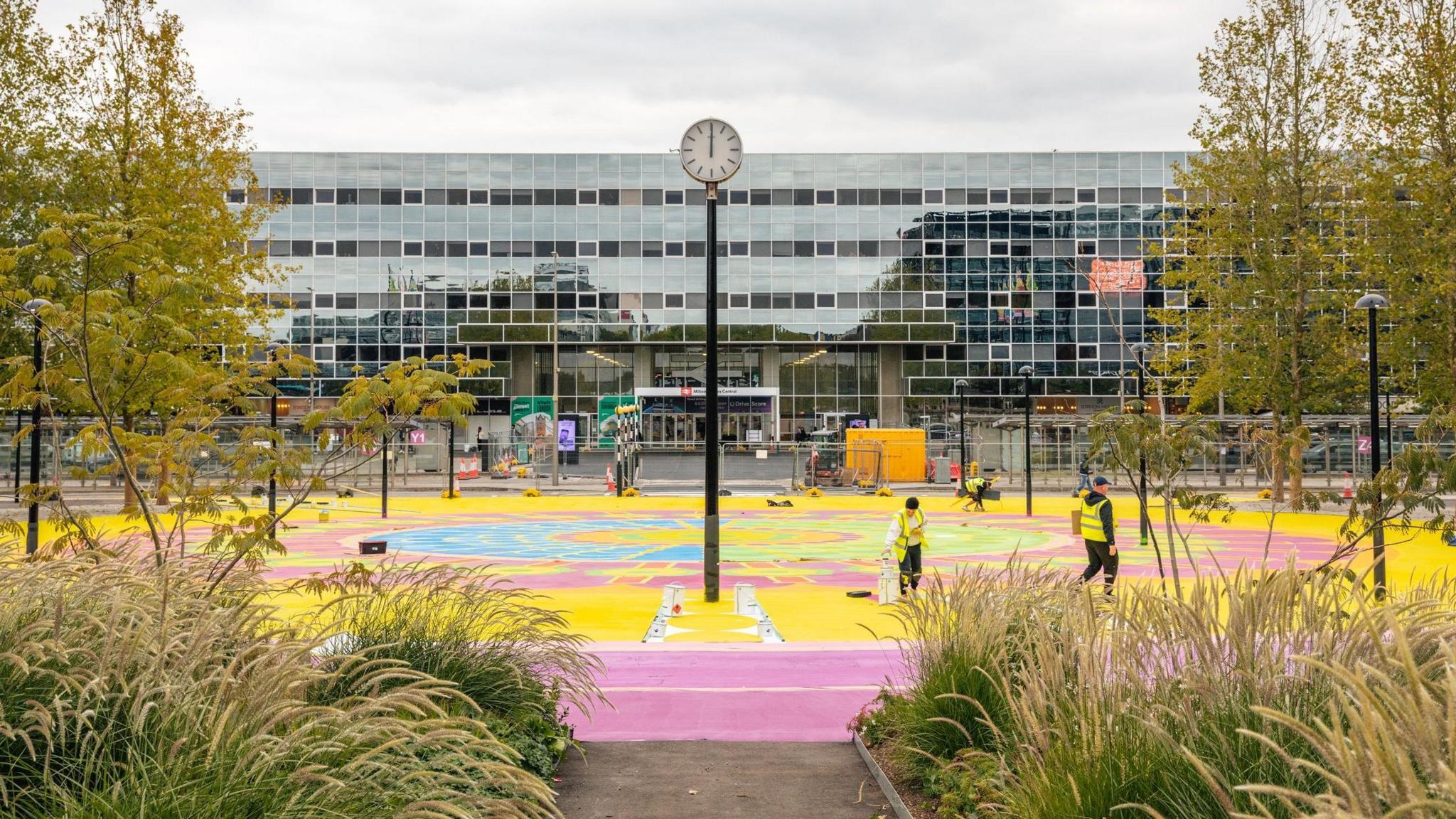 MK station building, which is wide and shiny and reflects some of the trees opposite it. The station forecourt displays the artwork: a large circle on the floor coloured bright pink, yellow and blue. A tall black pole with a clock on top is in the  the circle, and in the centre of the photo. Shrubs and trees surround the circle. Some painter in high vis jackets are working on the painting.