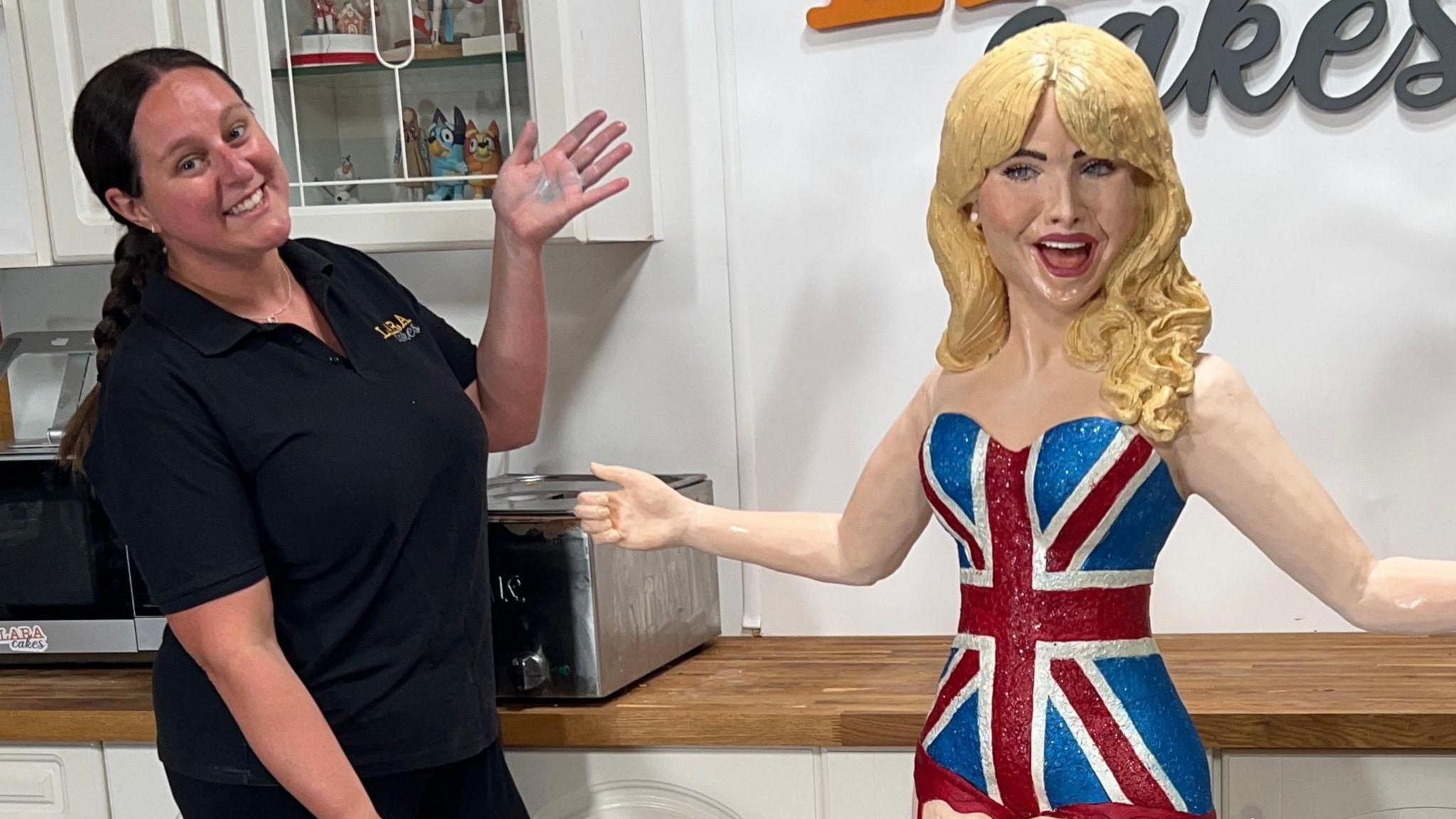 A cake version of Sabrina Carpenter. Her mouth is open and her blonde hair is covered in icing. She is wearing a Union Jack dress. Baker Lara is to the left of her gesturing