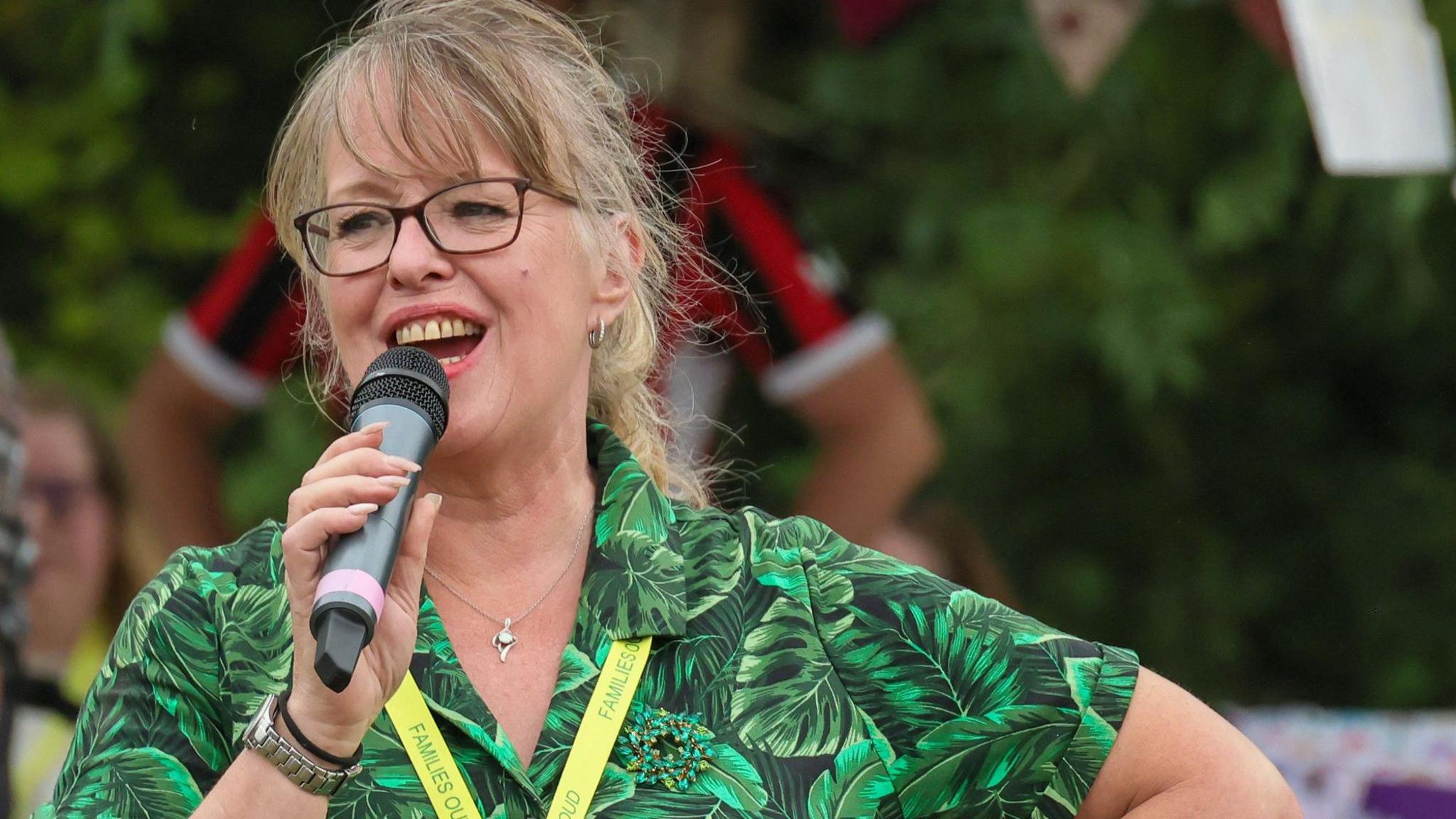 Tanya, wearing glasses and a green dress in a pattern with lots of large leaves, speaks into a microphone at an outdoor event.
