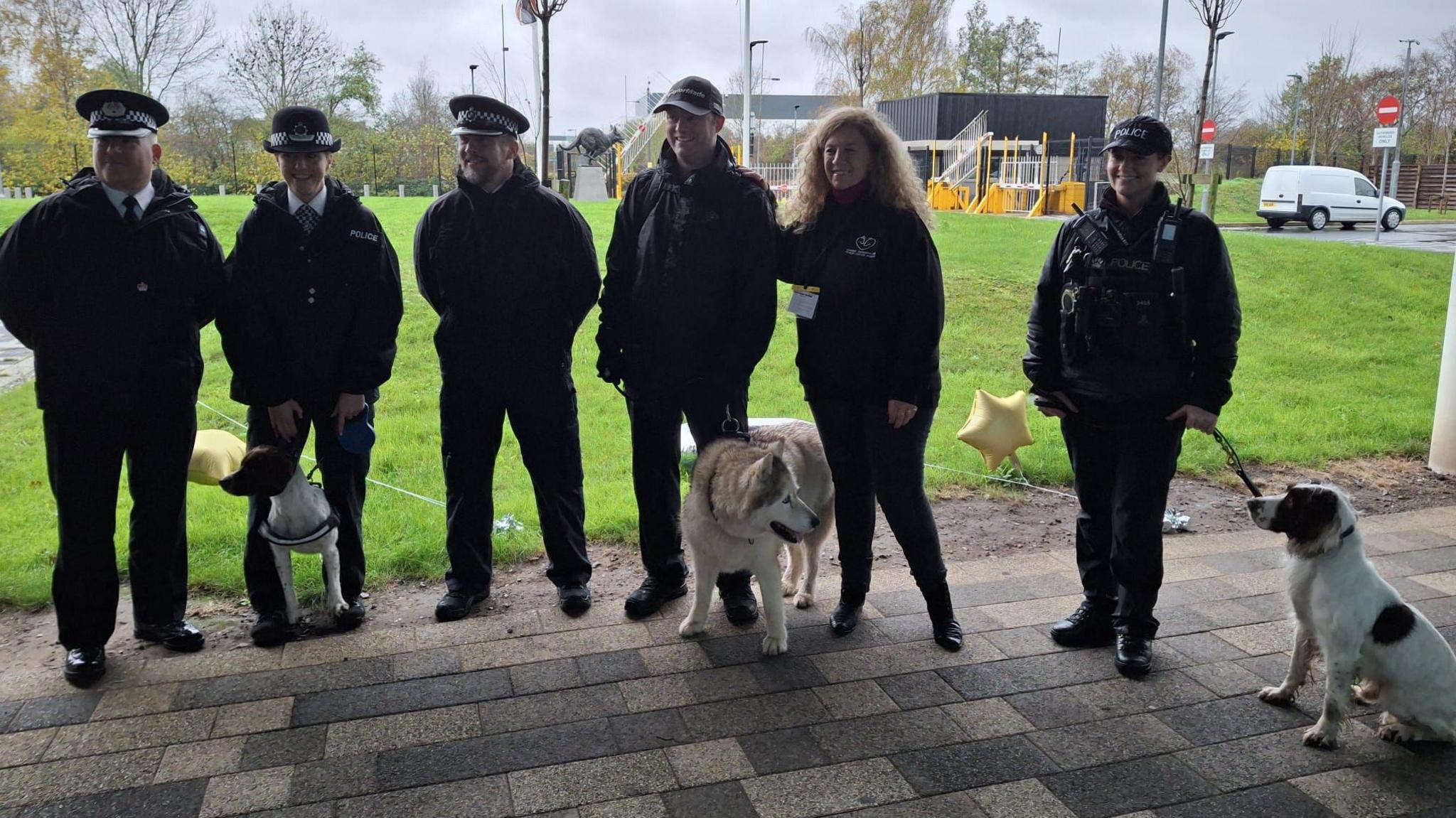 A group of police officers standing in a line with Lady Bathurst, who is wearing a waterproof jacket. Three of the officers are holding dogs on leads, including a husky and two spaniel type breeds.
