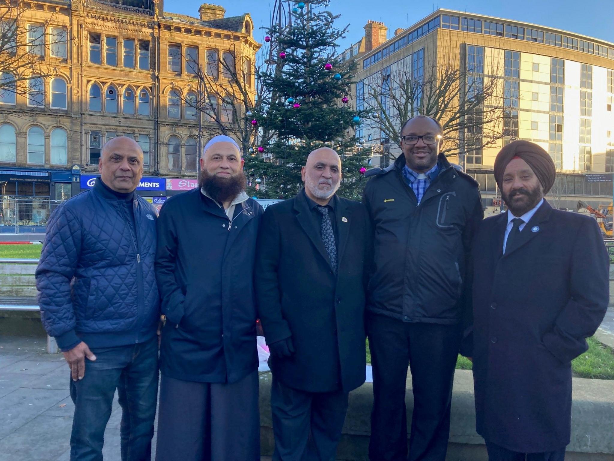 Five men wearing dark clothes standing in front of a fir tree with pink baubles