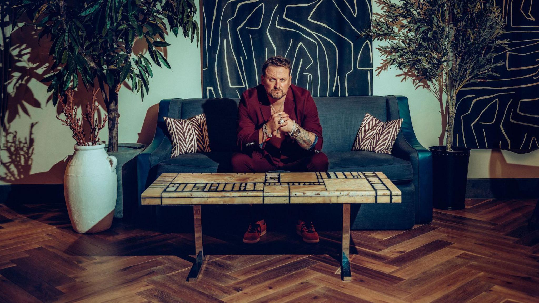 Vince Freeman in a red suit sitting on a blue sofa with his hands held together