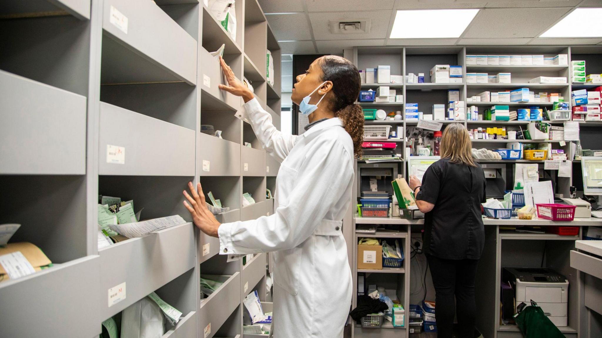 A woman in a white lab coat in a pharmacy wearing a mask. There are numerous shelves with packs of medication on them. 