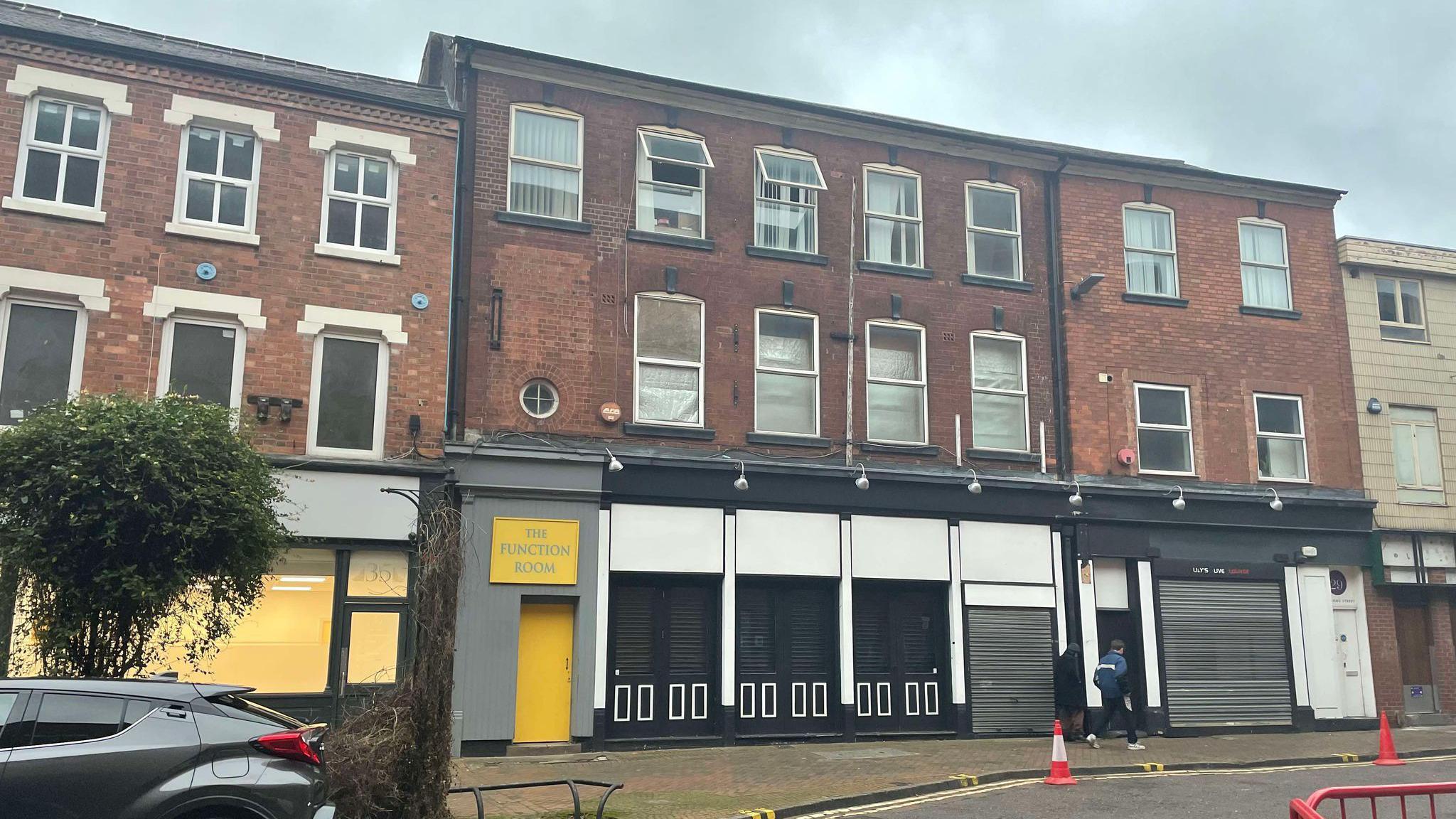 A shuttered up three storey building on a city centre street, with a yellow side door with a sign sating The Function Room and a sign on the right hand side of the shop front saying Lily's Live Lounge