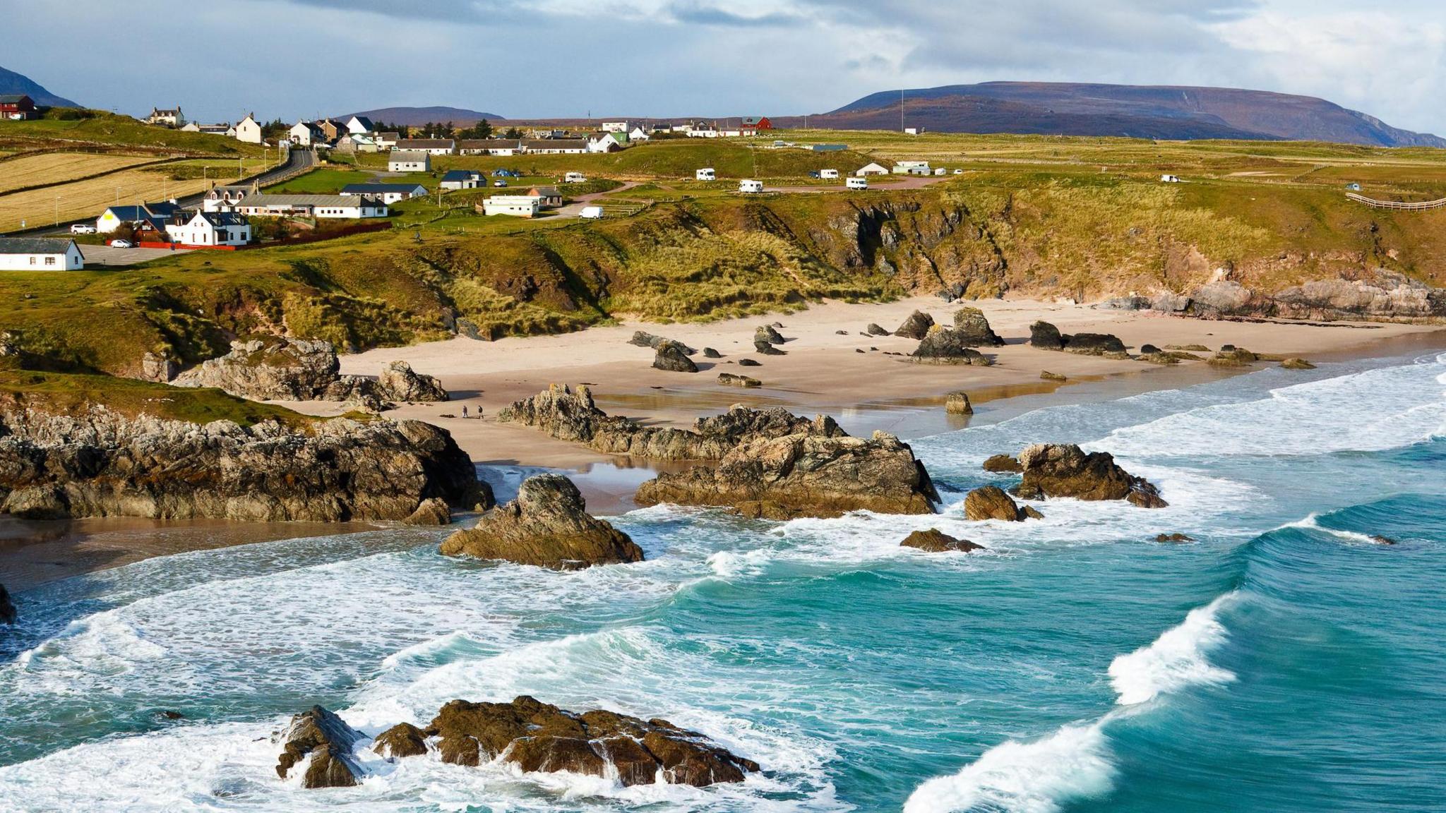 White walled properties dot an area of land near small cliffs above a beach. Waves roll in from the sea, which is a turquoise colour. There are rocky outcrops on the beach and in the sea.