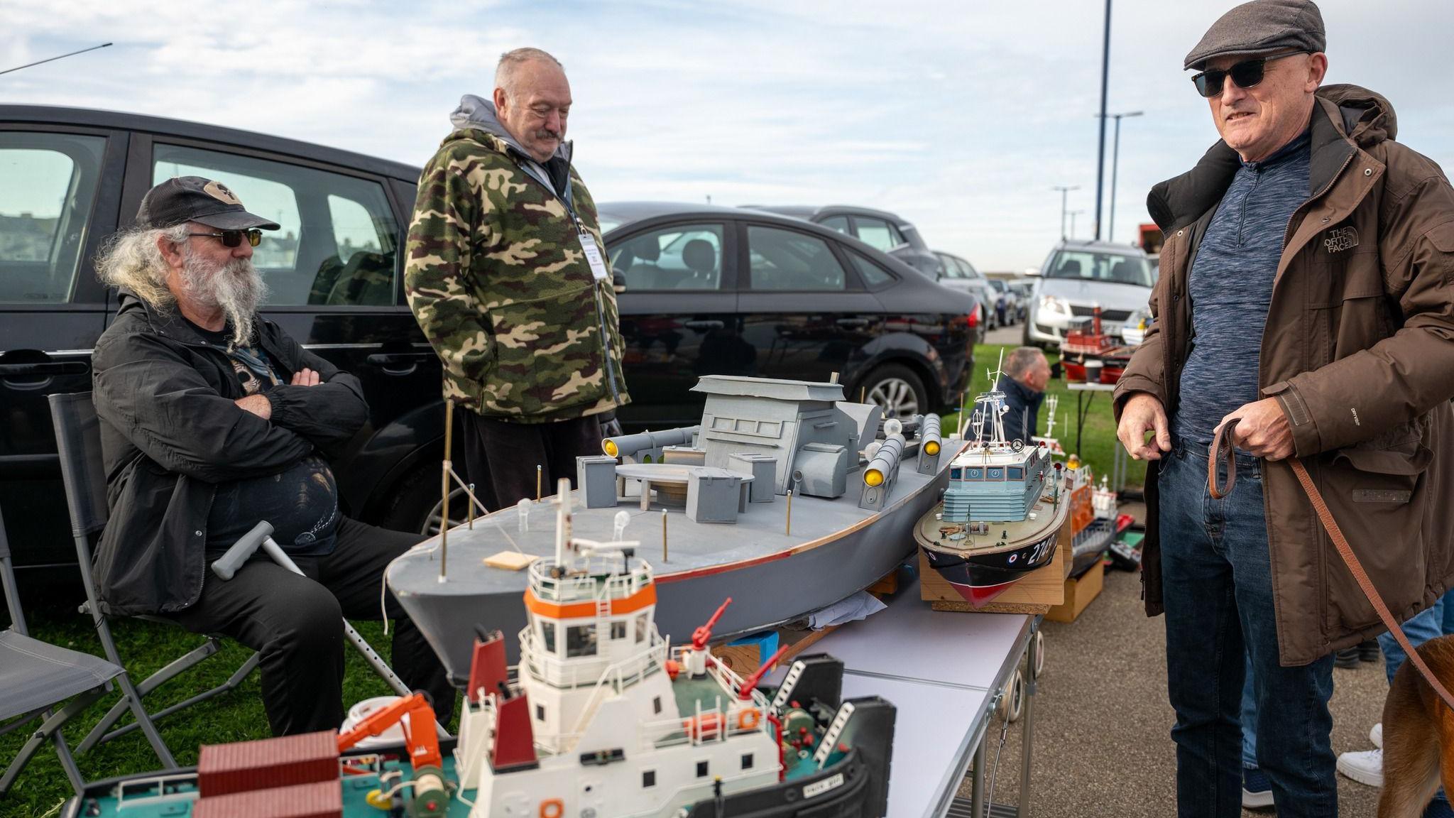Three colourful model boats, including a large grey one, are place on a table. Three men are looking at the boats. There are parked cars behind them.