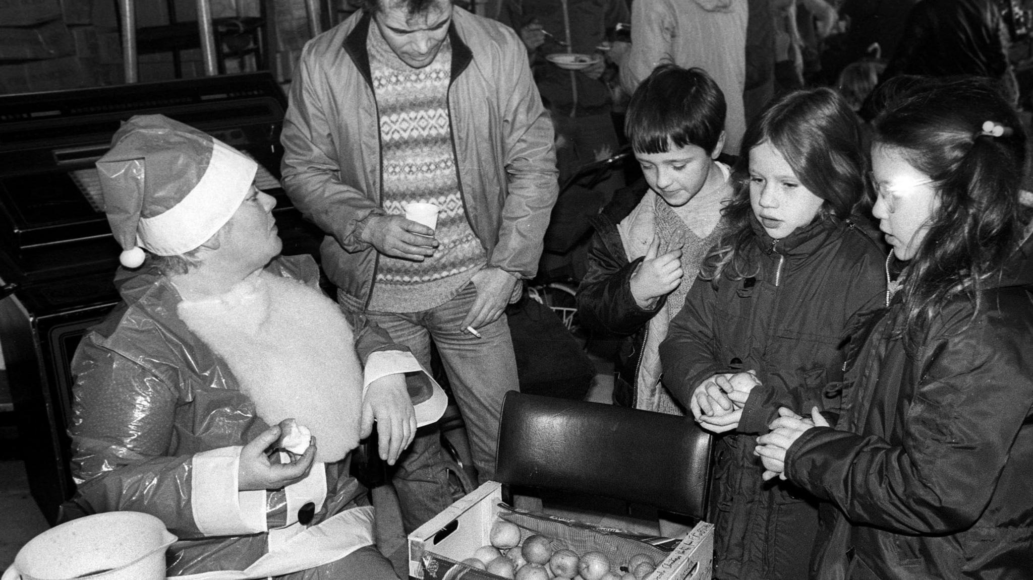 A person dressed as Father Christmas talks to a young man and three children. A box of fruit sits on a leather chair between the party.