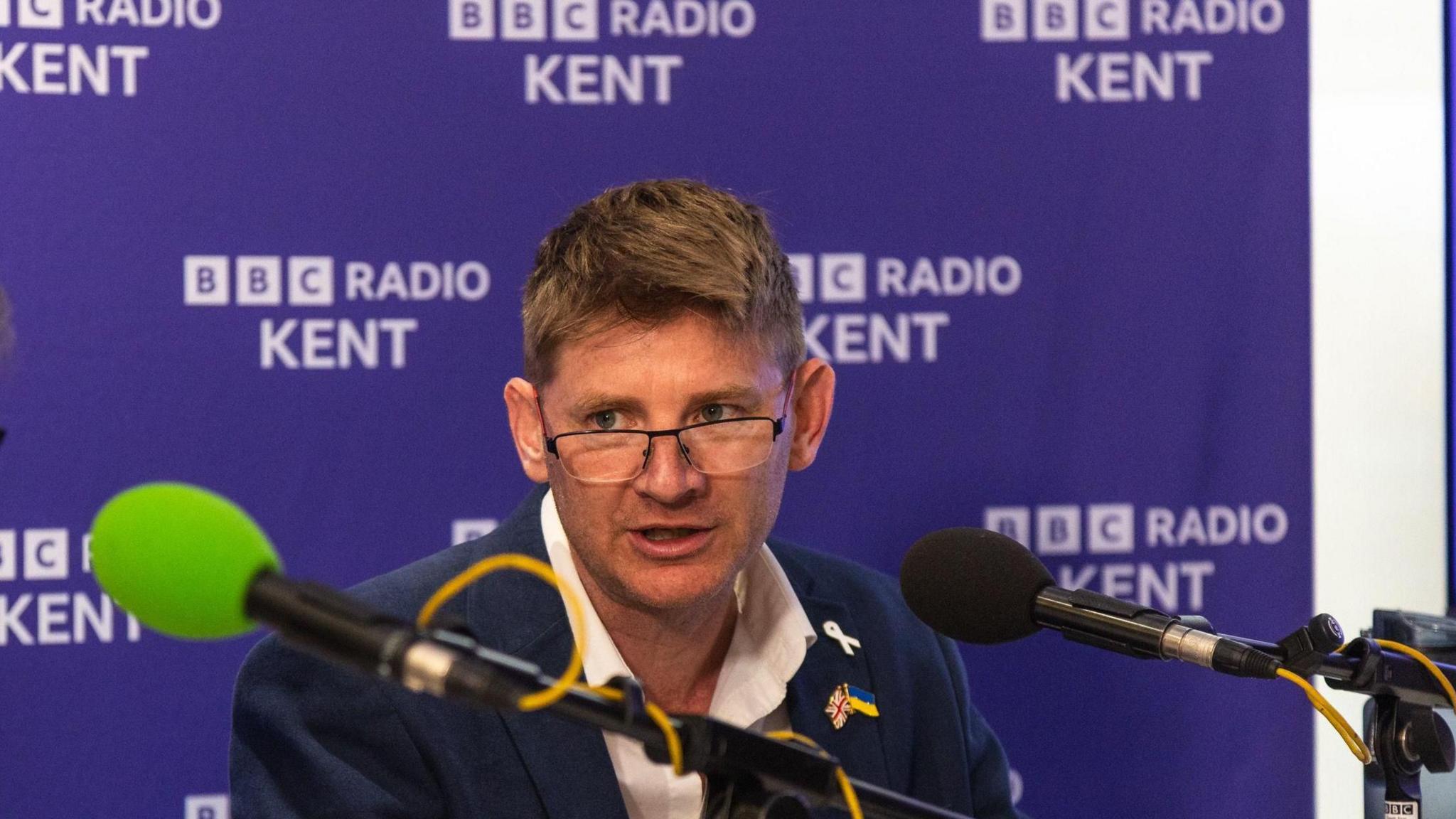 A man with glasses, wearing a white shirt and blazer. He has a badge of the combined flags of the UK and Ukraine pinned on his lapel. He is speaking into a microphone and sits in front of a BBC Radio Kent purple and white backdrop.