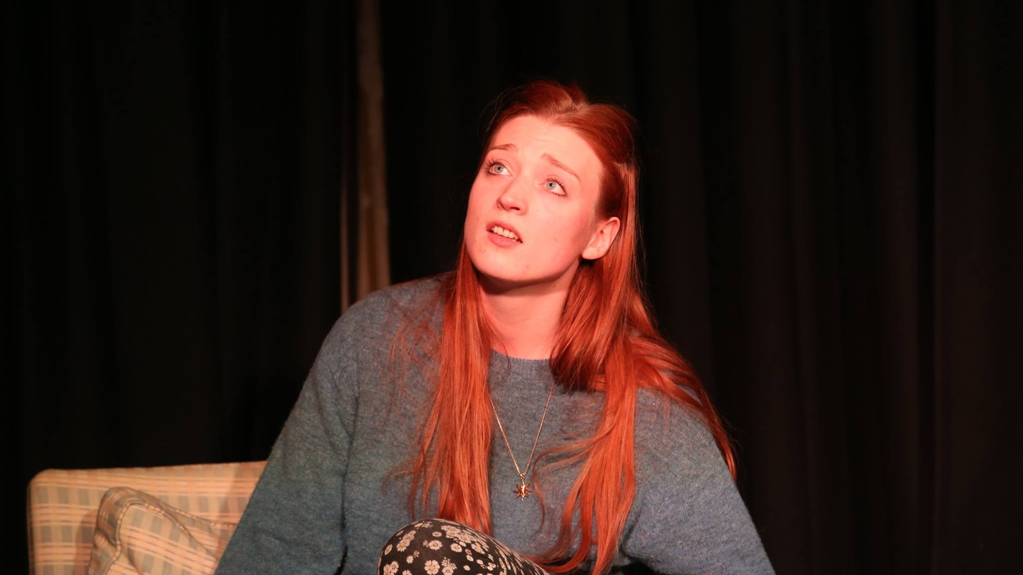 A young red-headed woman in a grey jumper sits on a chair on a black stage, looking into the distance.