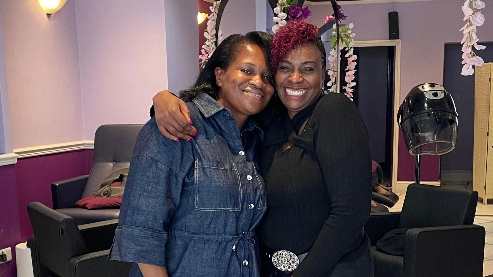 Wilma Burke smiles brightly alongside her client Katherine Kidd at Pride Hair and Beauty Salon. Wilma wears a long black fitted shirt with a silver-buckled belt, black trousers and has burgundy hair. Katherine, in a denim shirt tied at the waist, has freshly styled shoulder-length black hair. Both women look directly at the camera, smiling.