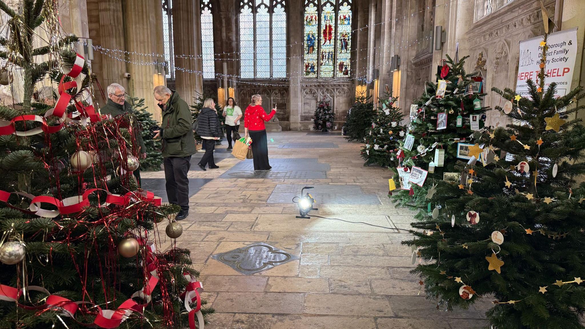 Green Christmas trees with decorations - the trees on the left ahve red decorations with golden balls and the trees on the left have mostly yellow and white decorations including cards. They are placed inside the cathedral building with stained glass windows in the background and people walking around the display.