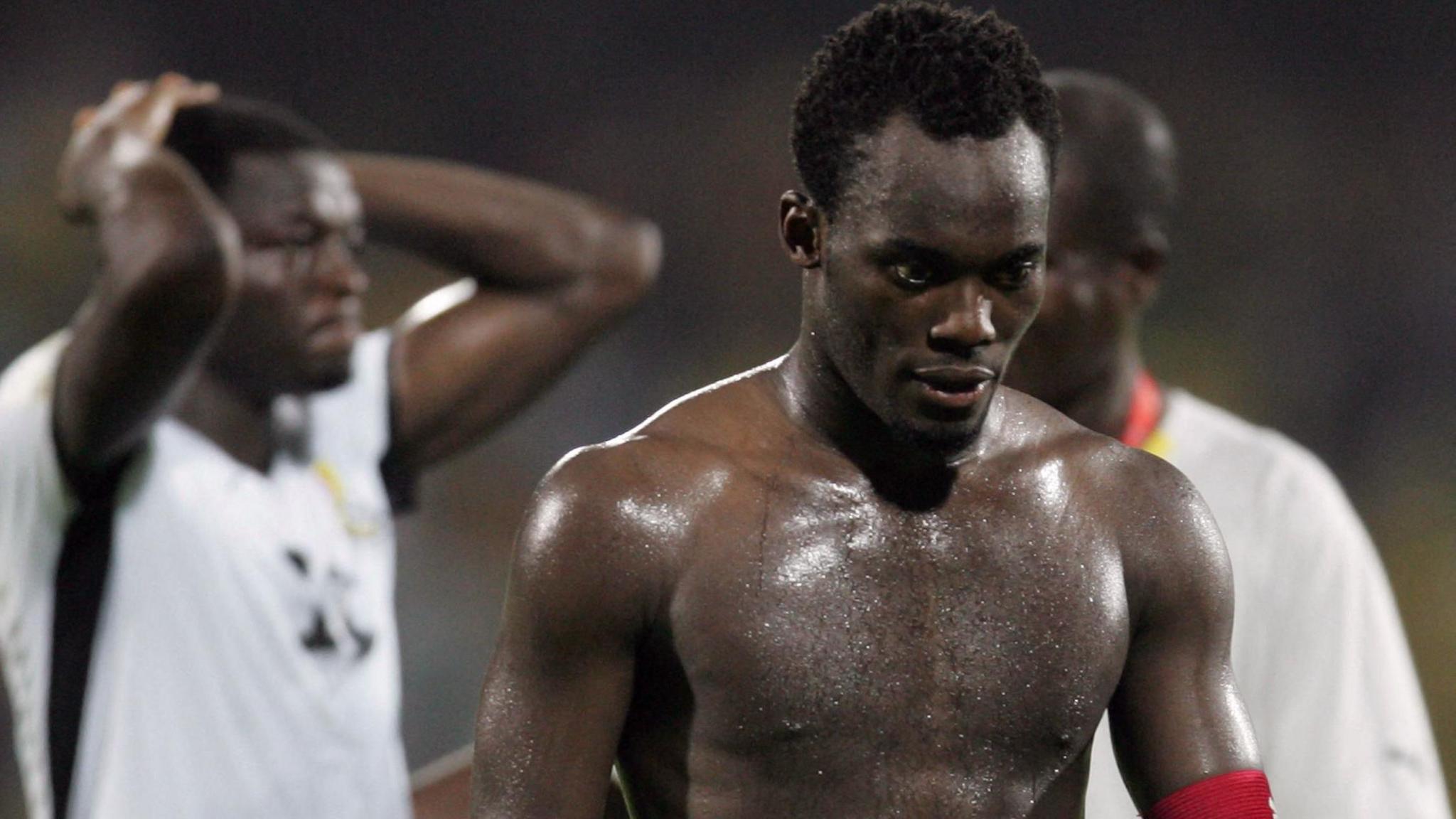 A topless Michael Essien, with just a red captain's armband on his left bicep, is visible from waist up as he trudges off the pitch after Ghana lost a match at the 2008 Africa Cup of Nations. An out-of-focus Ghana team-mate can be seen in the background holding his hands to his head in disappointment, while another is obscured behind Essien