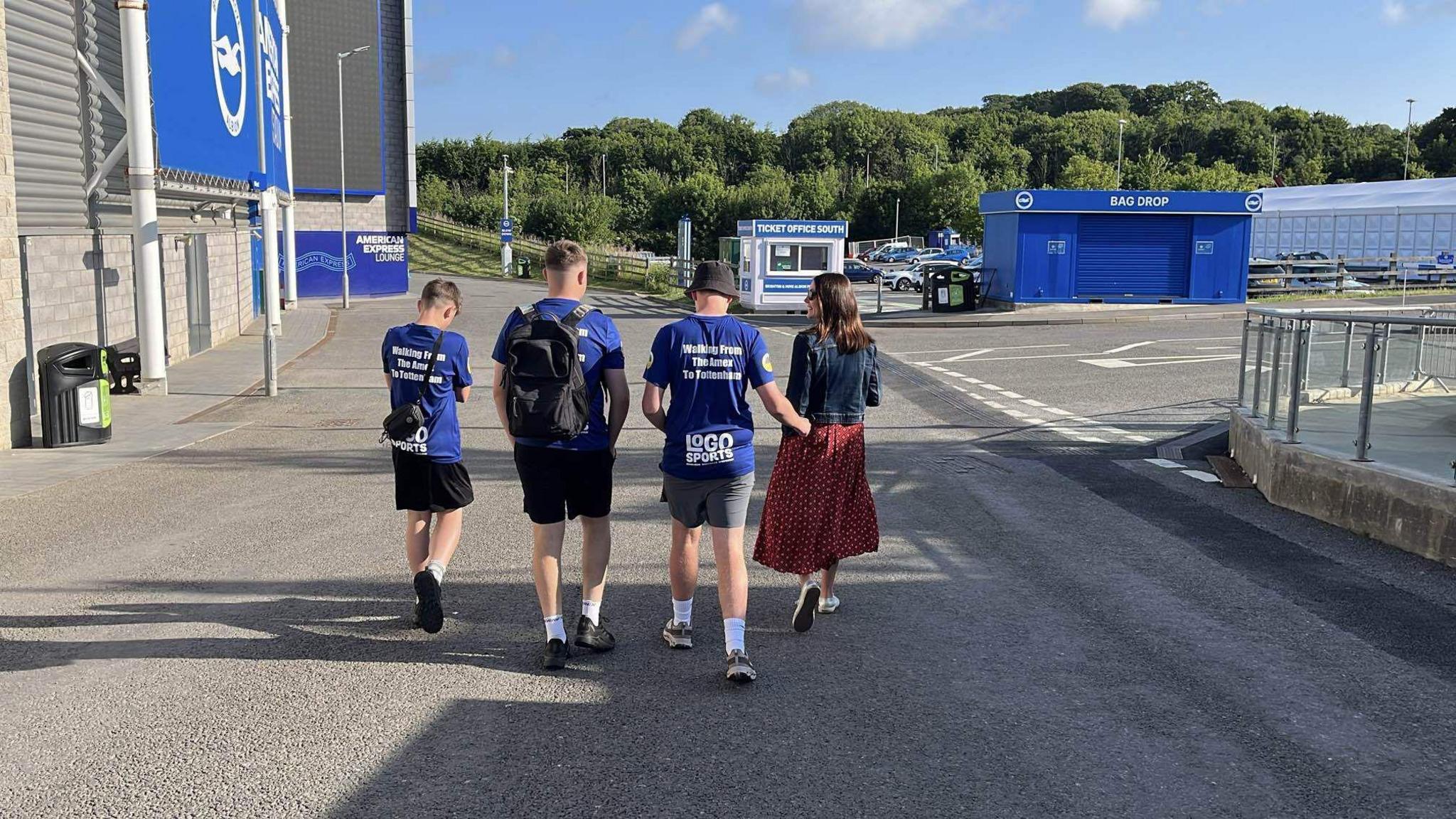 Three teenagers in matching blue shirts walking away from the camera outside the Amex stadium in Brighton. They are accompanied by a woman in a denim jacket and red skirt.