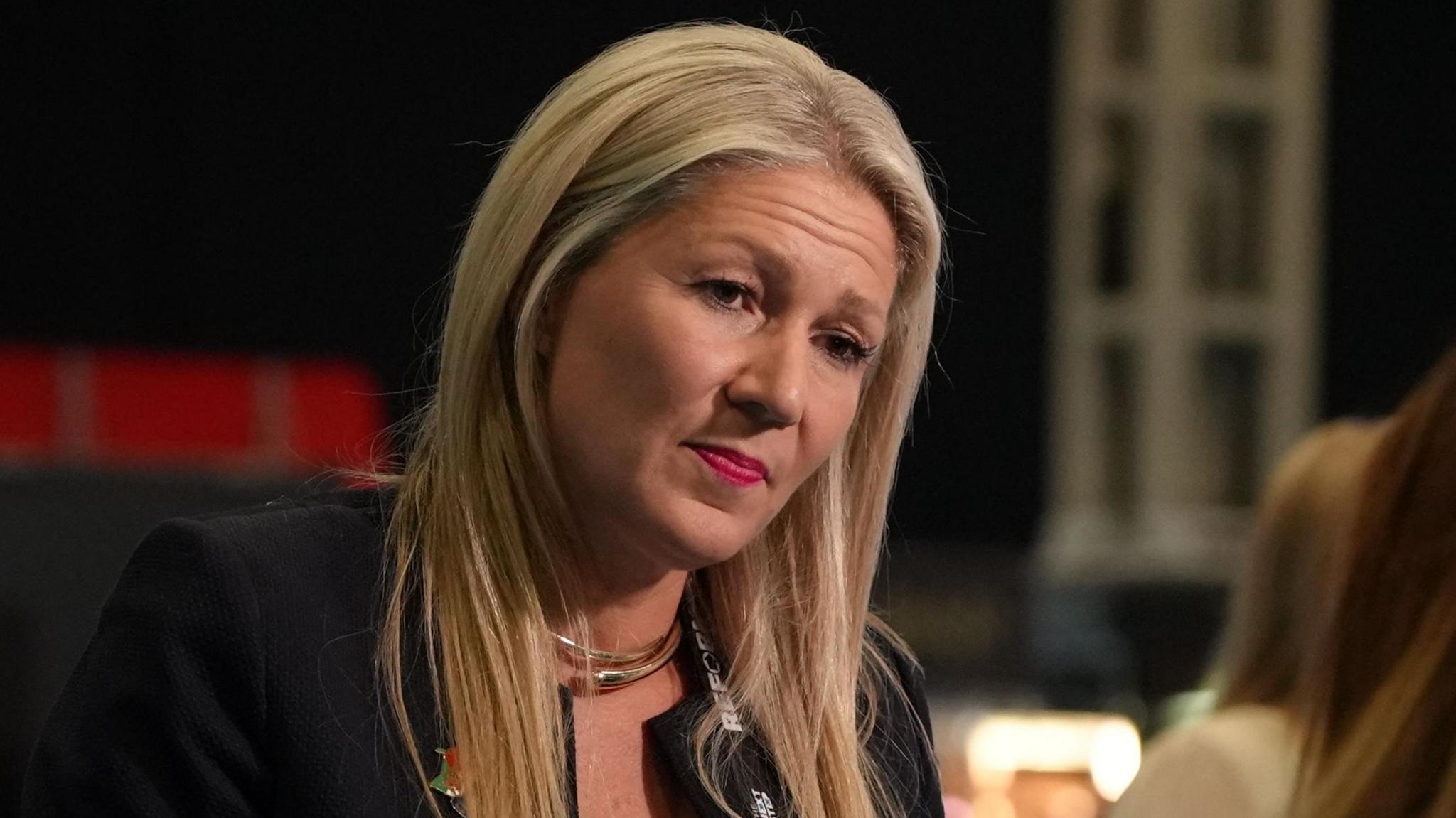 Laura Anne Jones wearing a black top, in conversation with a person slightly off camera. She has a black backdrop behind her and is wearing a Reform party lanyard.