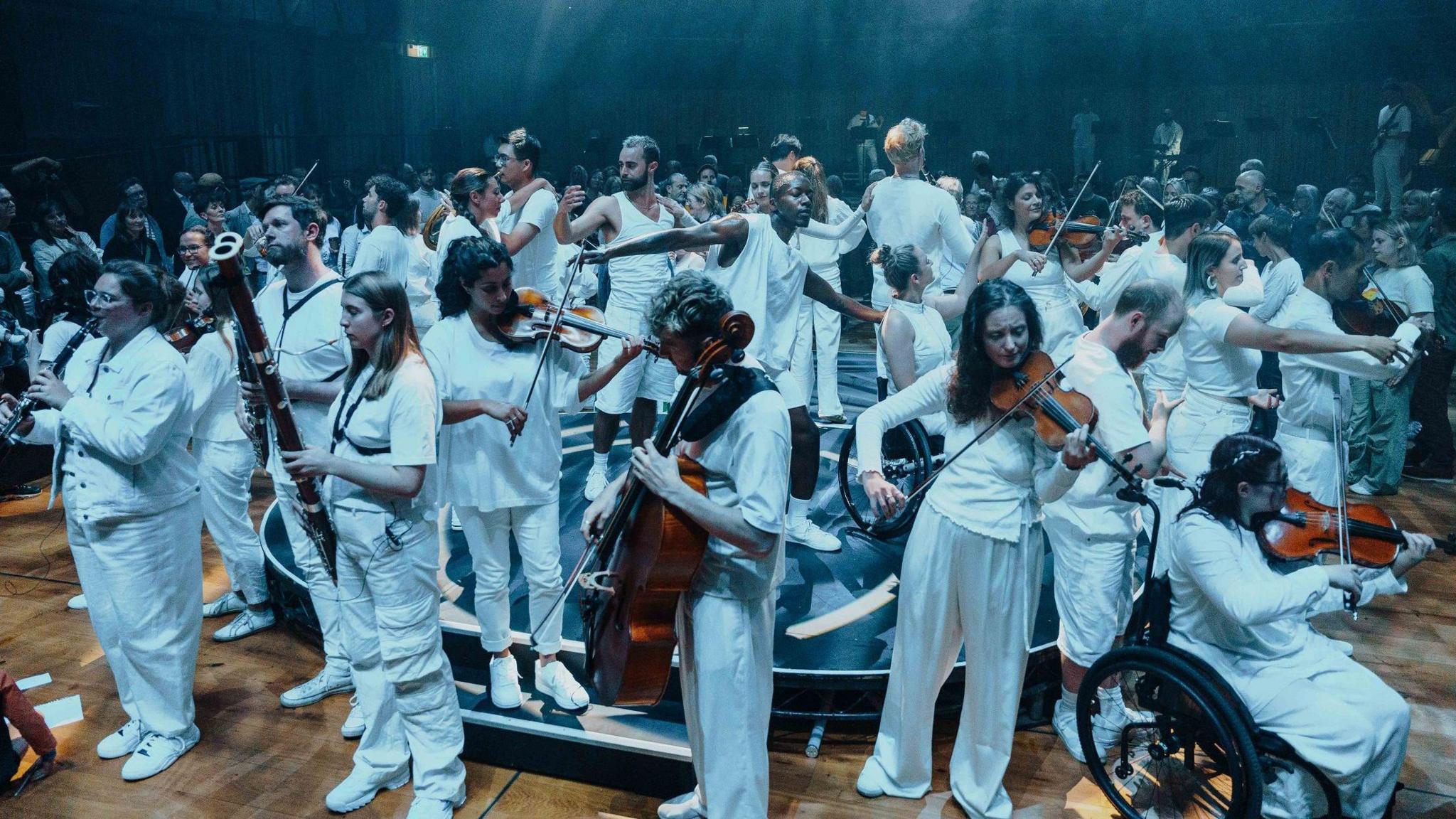 Dozens of musicians, all dressed in white, perform at the centre of the auditorium at Bristol Beacon as part of the BBC Proms weekend in the city. The musicians, members of Paraorchestra, are playing a variety of instruments including violins, cellos and oboes