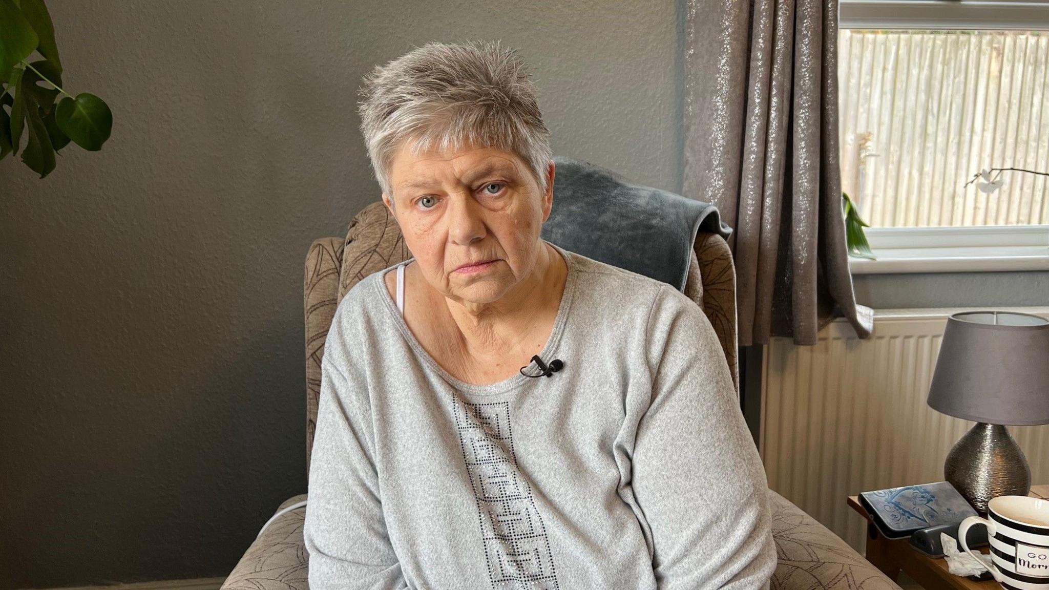 A woman in her sixties with short grey hair is looking directly into camera. She is wearing a grey jumper and sitting on a grey lounge chair.