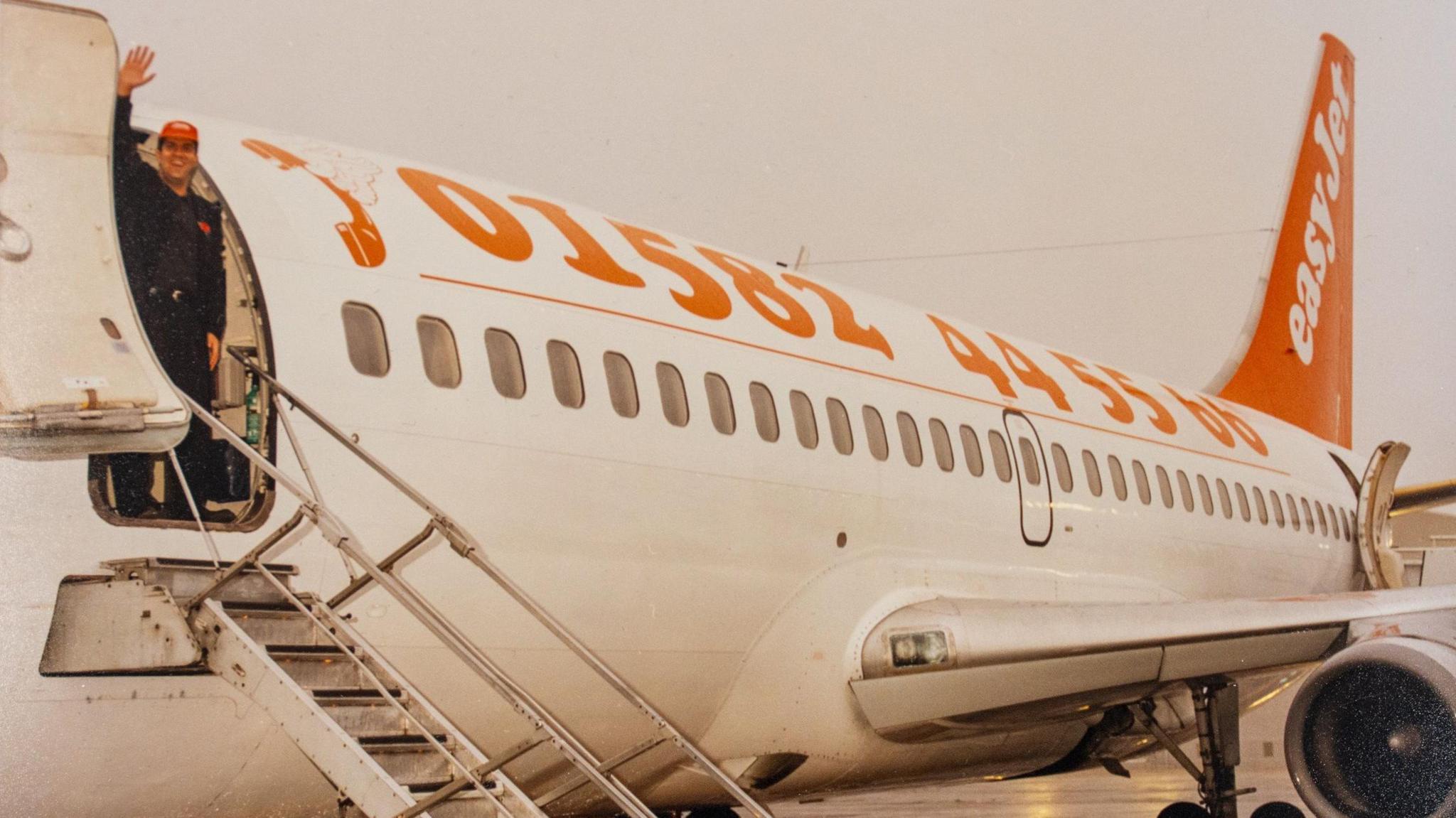 Stelios stands at the top of the plane's steps waving at the camera. He wears an orange Easyjet baseball cap. The side of the plane has the Luton Airport phone number emblazoned in orange against a white plane. Is says 01582 445566. The tail of the plane is orange and has easyjet written in white.