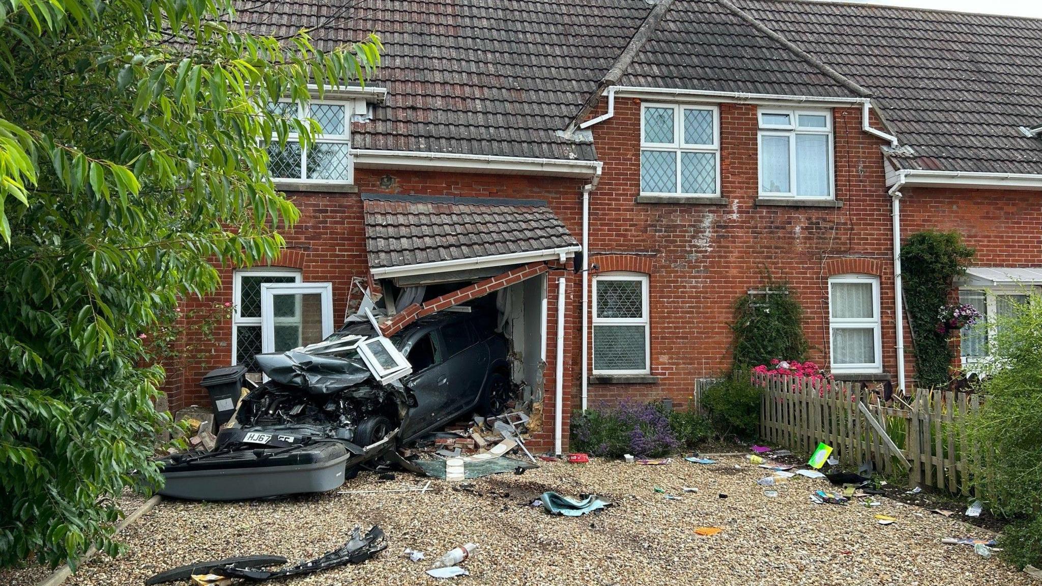 The front of a smashed up grey car which can be seen with its rear crashed into the front of a house. The door of the house is destroyed and the porch of the house is hanging down. There is a lot of debris on the driveway of the house. 