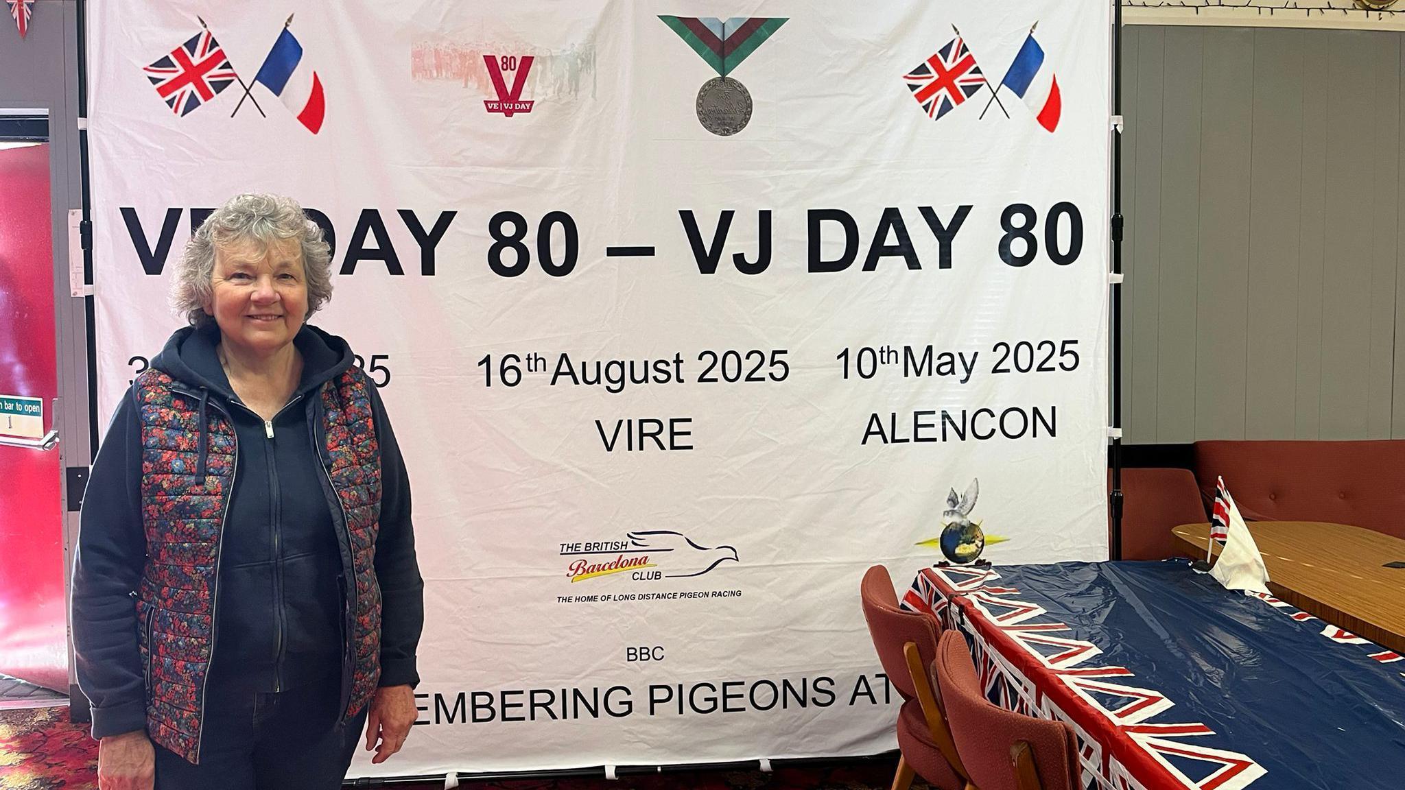 Jill Rogers stands in a room in front of a poster which reads "VE Day 80 - VJ Day 80" and lists dates of three pigeon races starting in France. She has short, grey hair and wears a gilet over a dark blue hoodie. The room also has a table with a Union Jack-decorated covering.