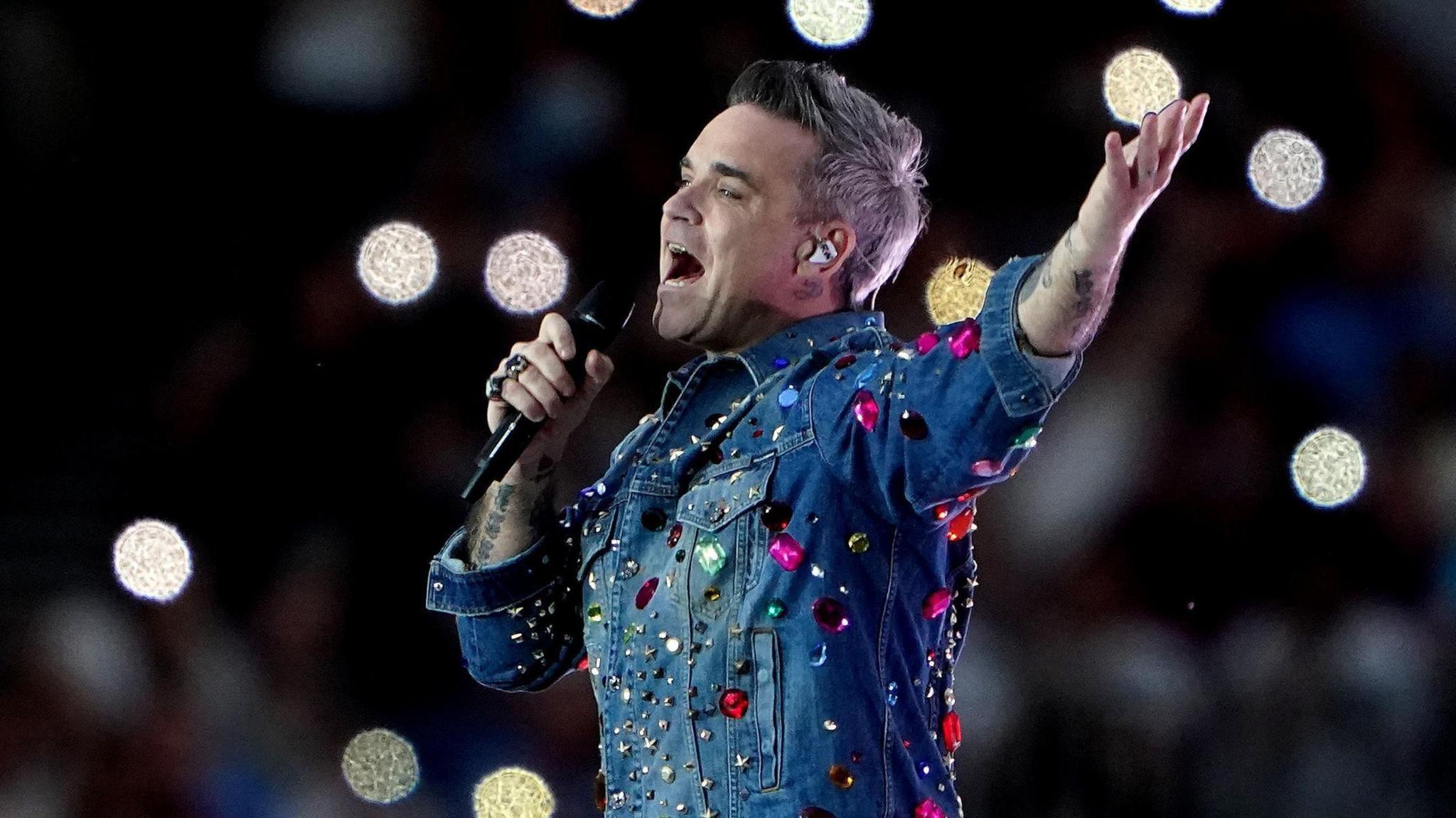Robbie Williams wearing a denim shirt with jewels stuck on it. He is pictured side-on while performing at a concert. He is singing into a microphone held in his right hand while his left arm is raised in the air.
