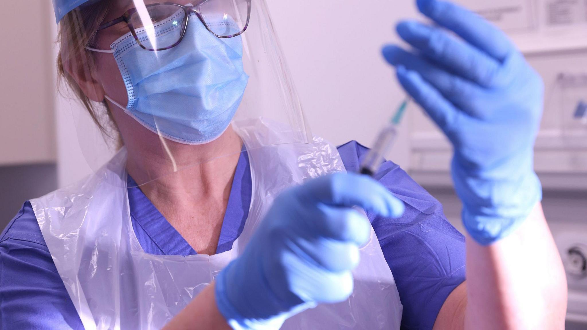The picture shows a nurse wearing blue rubber gloves, a blue mask and a clear visor. She is filling a syringe with the Covid vaccine