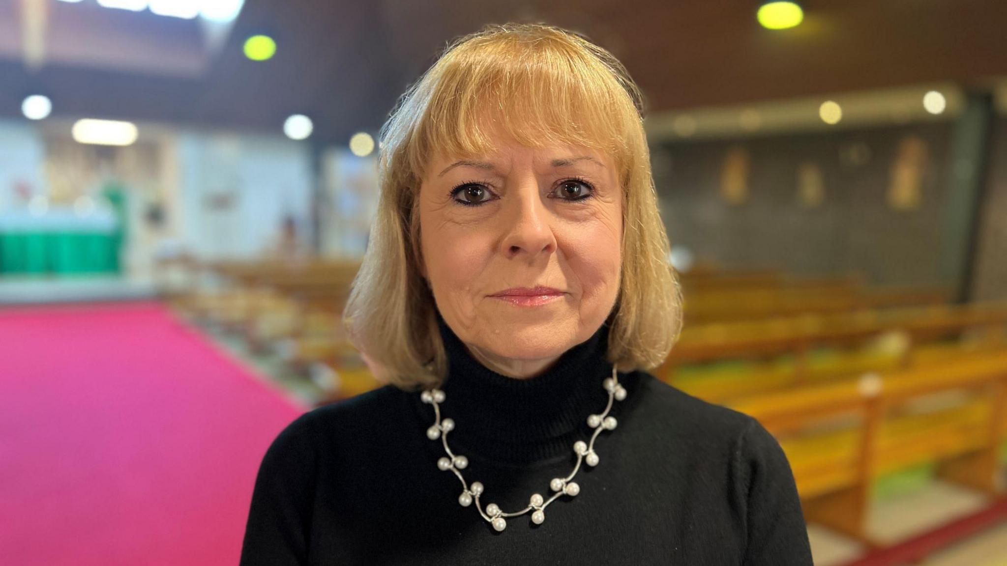 Woman with blond hair wearing a black polo neck jumper stands in an empty church.