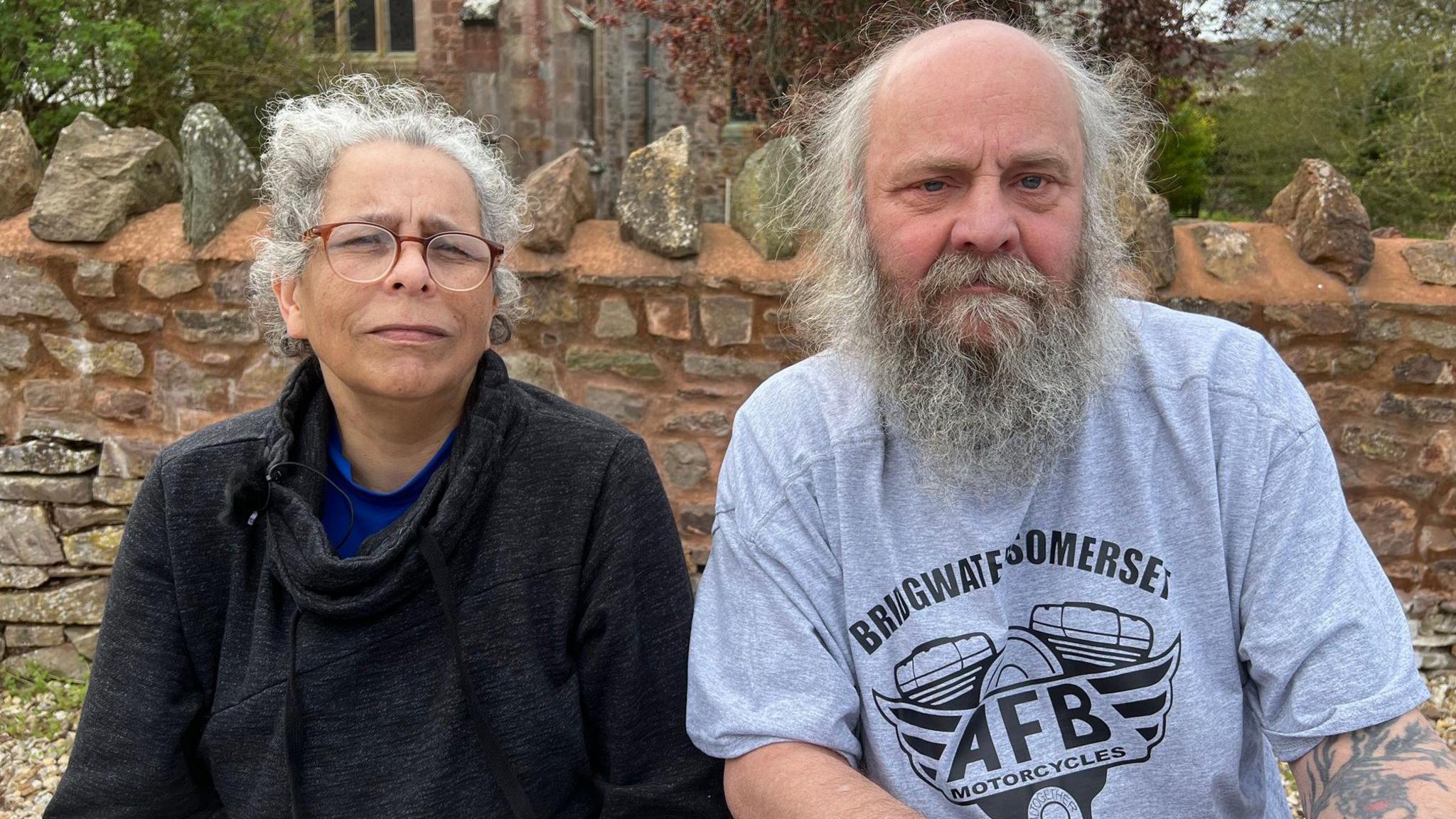 Two older people - a man and a woman - sit on a bench in front of a brick wall. The woman is in her 50s and has curly grey hair, and is wearing a black turtleneck and red-rimmed glasses. The man is in his 60s. He has a mostly bald head with wispy grey hair and a long beard. He is wearing light blue T-shirt with a motif on it and text that reads "Bridgwater, Somerset".