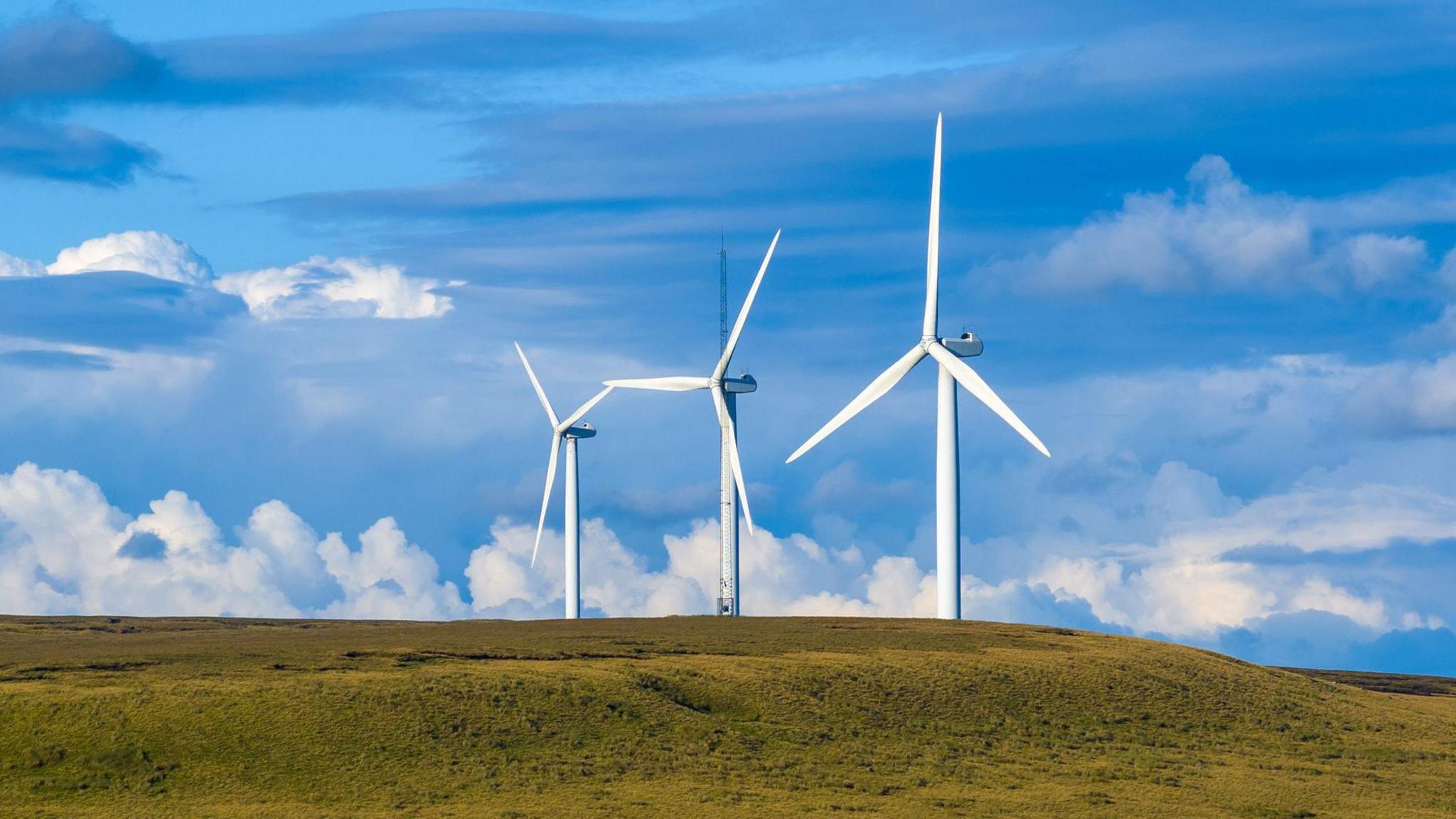 Three white wind turbines on a green headland set against a blue sky with fluffy clouds on a sunny day.