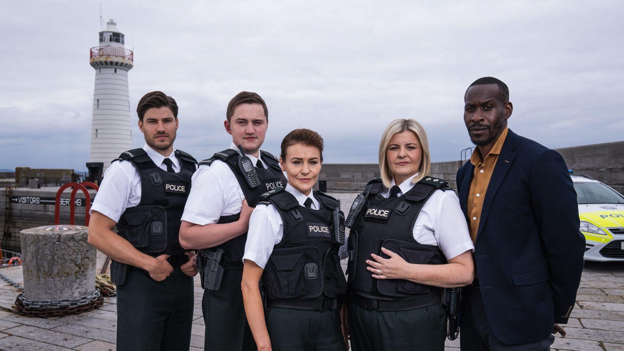 Hope Street actors Cameron Cuffe, Finnian Garbutt, Kerri Quinn, Tara Lynne O'Neill and Marcus Onilude in Port Devine.
They are standing in from of a lighthouse and a police car.
Four are wearing police uniforms of white short-sleeved shirts, dark trousers and a dark protection vests saying Police.
Marcus Onilude, stood at the far right, is wearing a dark blazer and a yellow shirt.