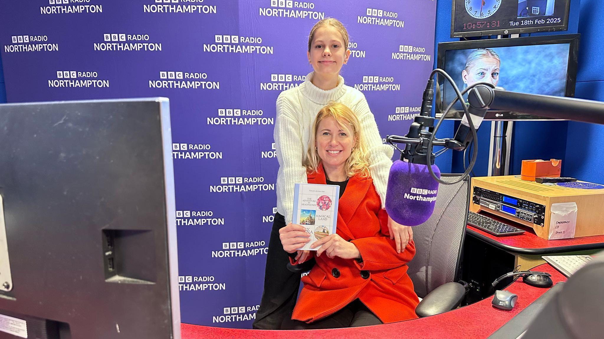 A woman in a red coat sits in a chair in a radio studio holding up a book. A girl in a white jumper stands behind the chair smiling at the camera. 