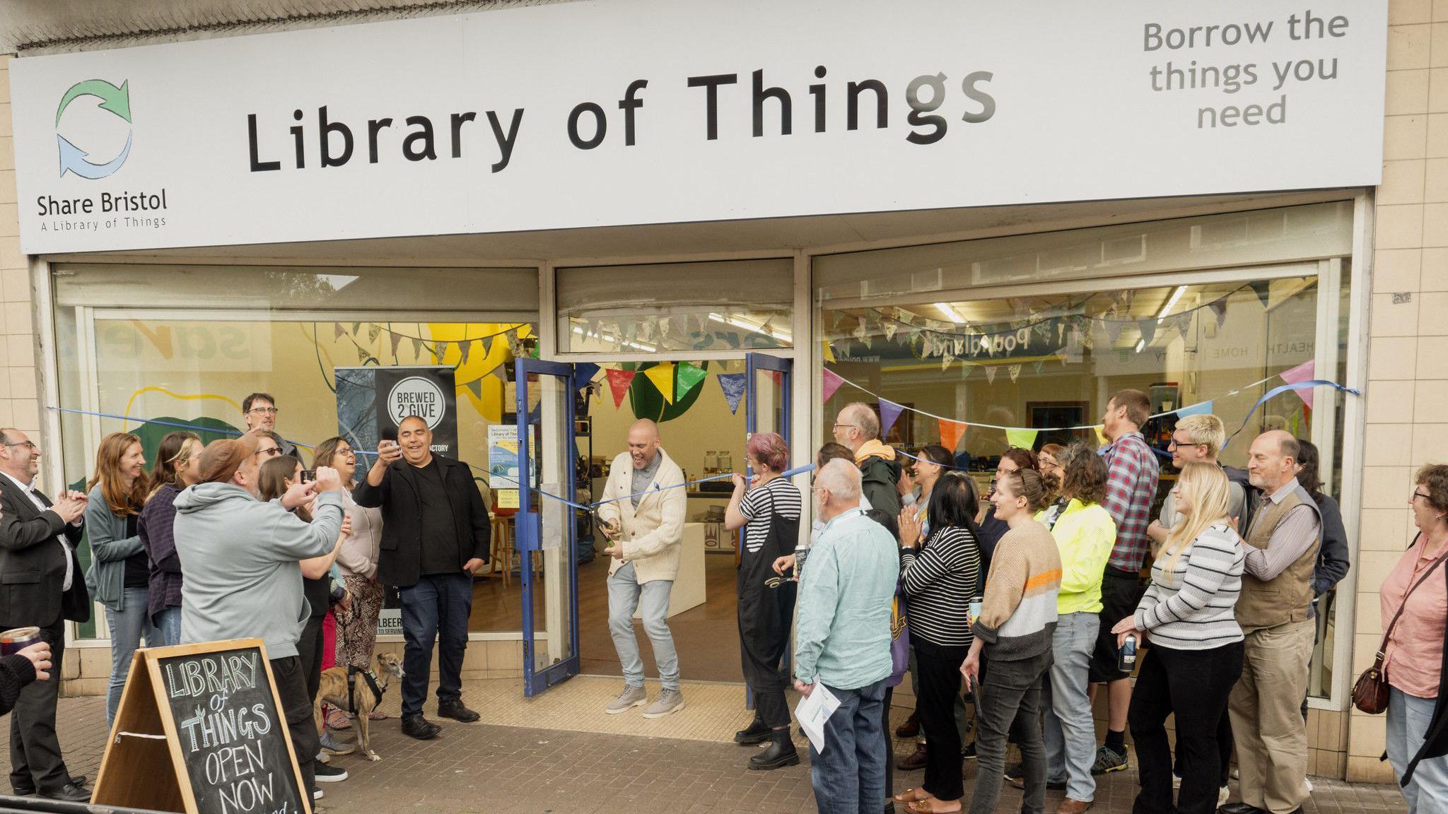 A crowd of people outside a shop with someone cutting a ribbon to open the building in the middle. "Library of things" is on the building's sign above the door.